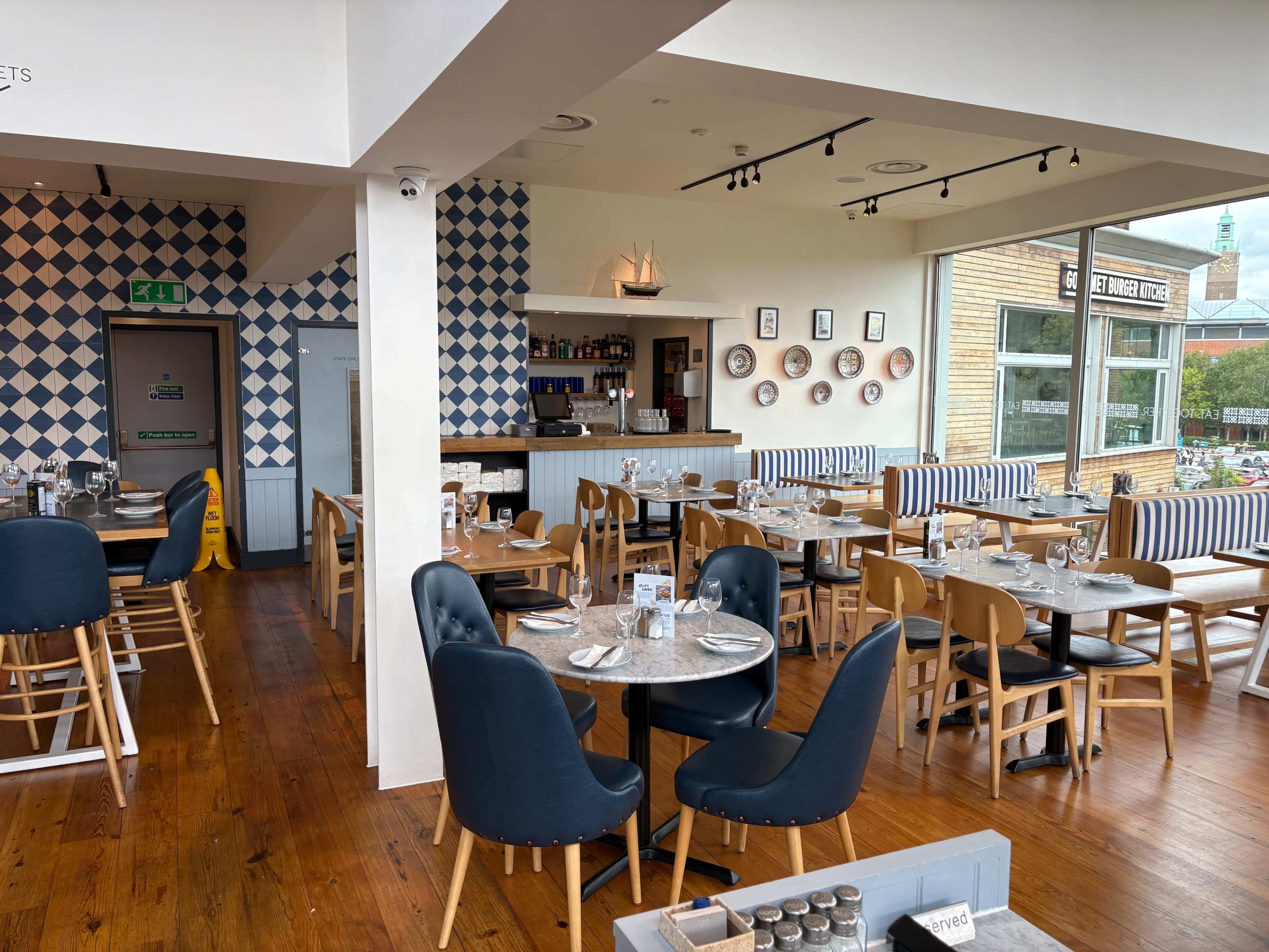 The image shows a restaurant interior with wooden floors, tables set with dinnerware, and blue checkered wall decorations.