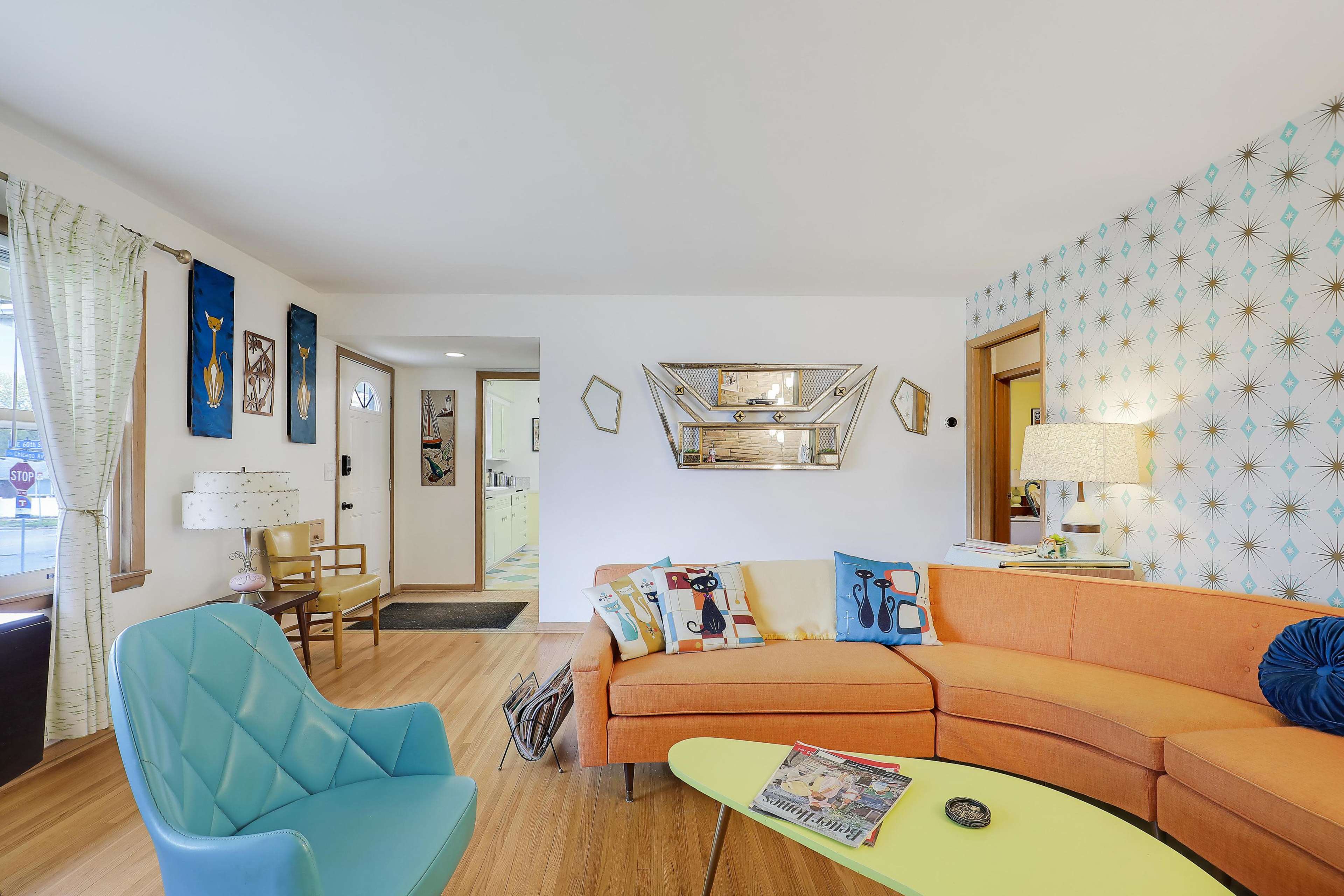 The image shows a brightly decorated living room with an orange sectional sofa, a blue accent chair, and a decorative mirror on the wall, complemented by geometric wallpaper and light-colored curtains.