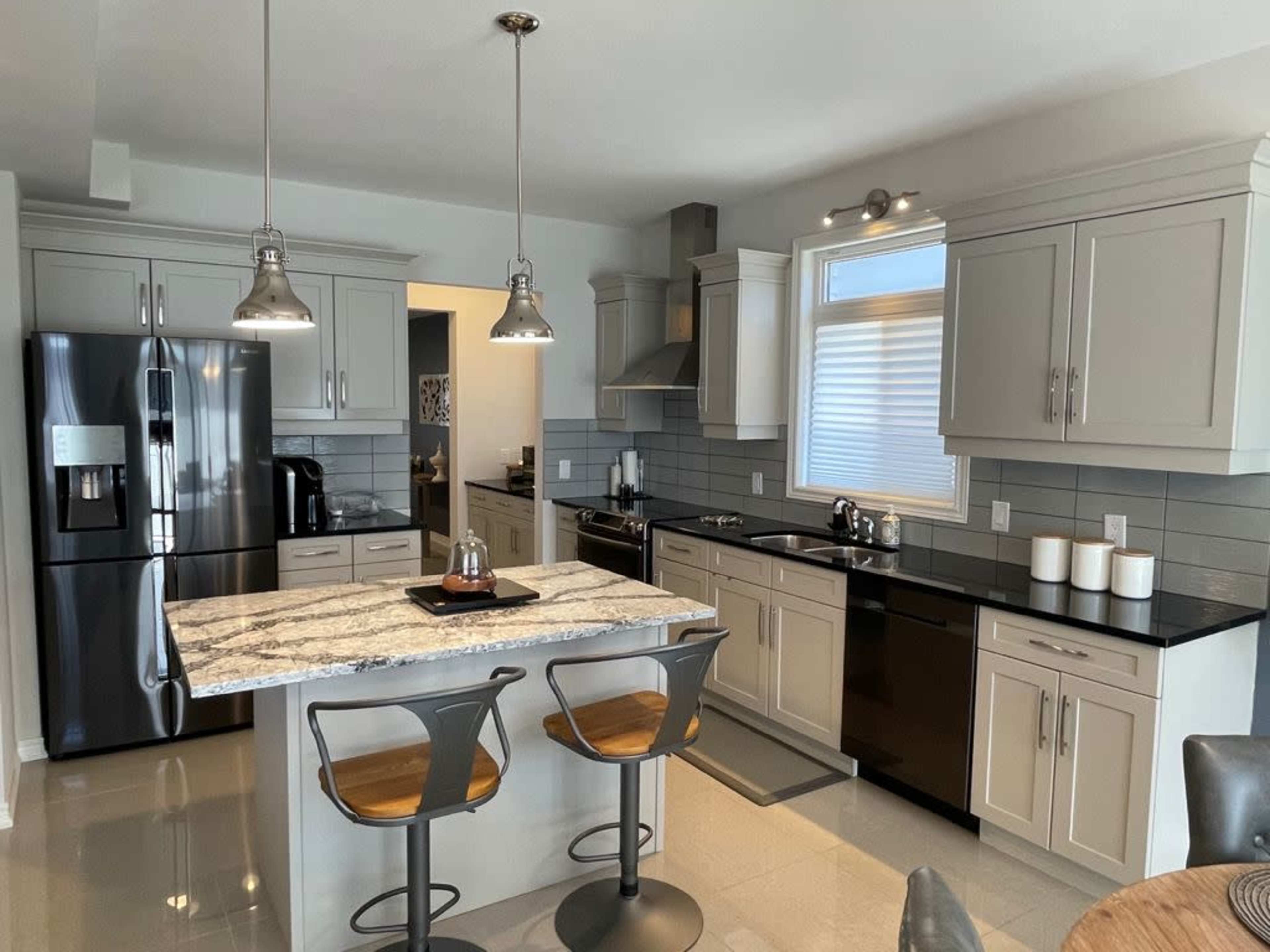 The image shows a modern kitchen featuring light gray cabinets, a large island with two bar stools, stainless steel appliances, and a black countertop.