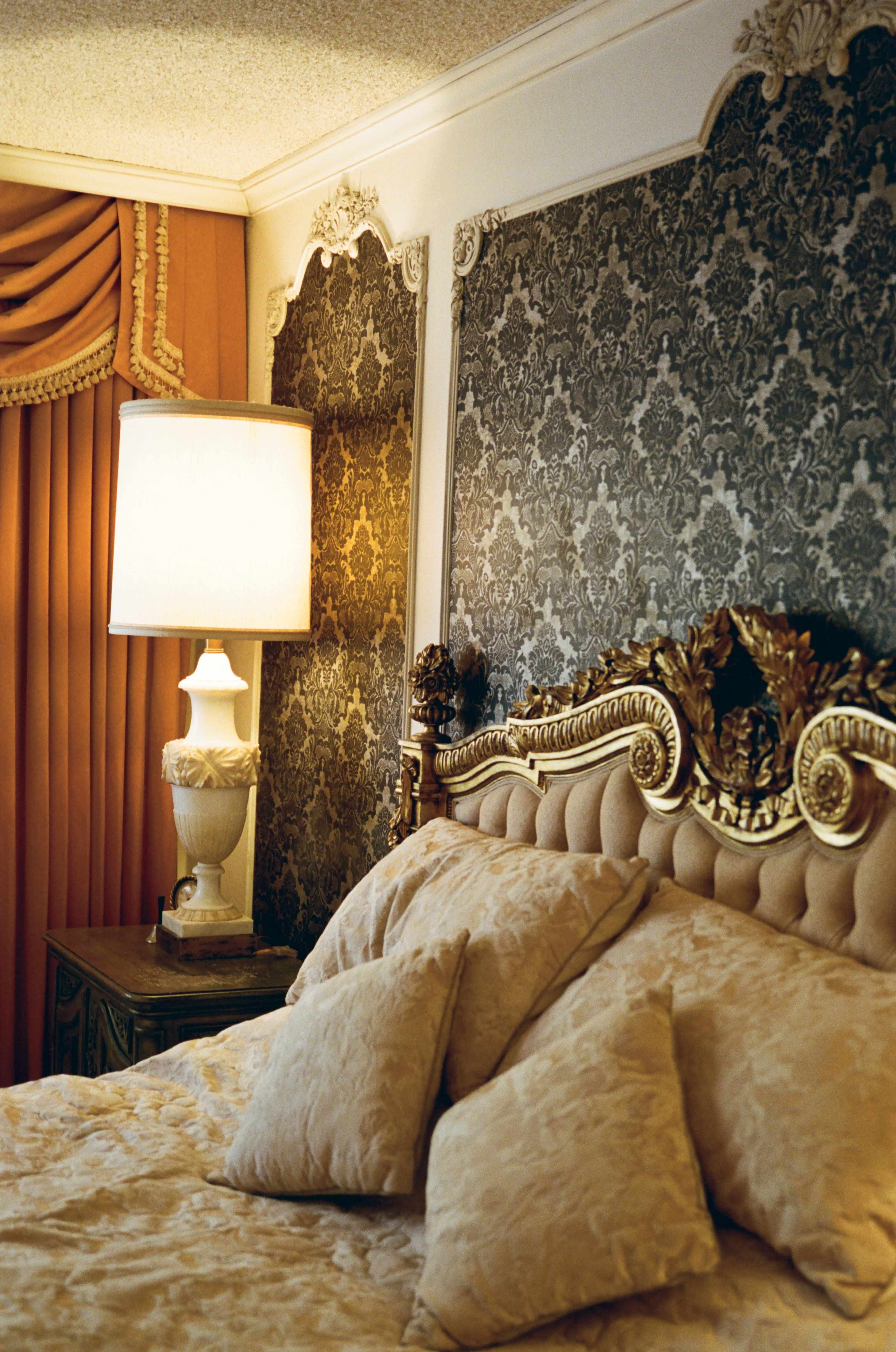 The image shows an ornate bedroom featuring a plush bed with decorative pillows, a vintage lamp, and patterned wallpaper.