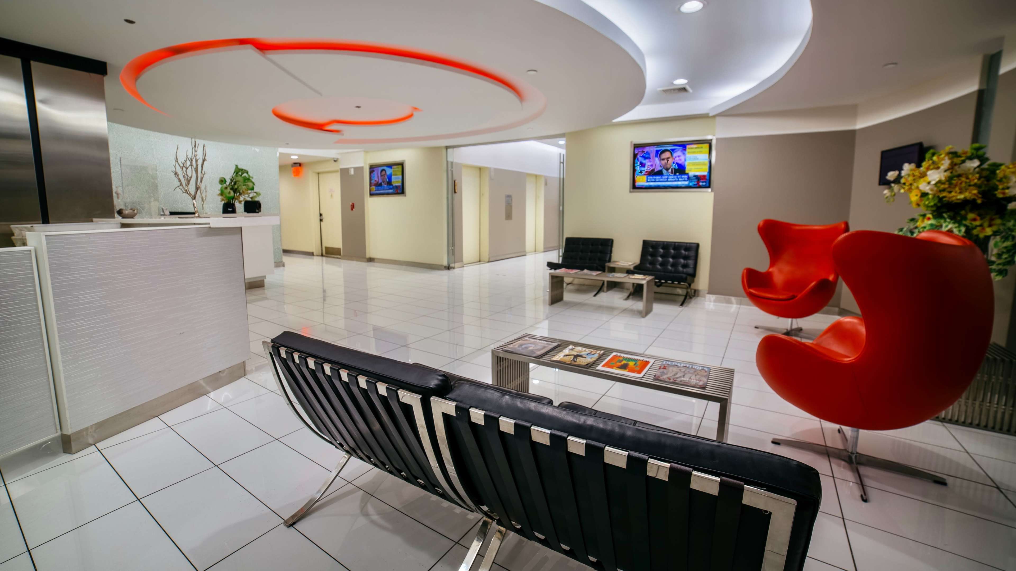 A modern lobby features sleek furniture, pendant lighting, and two flat-screen TVs mounted on the walls.