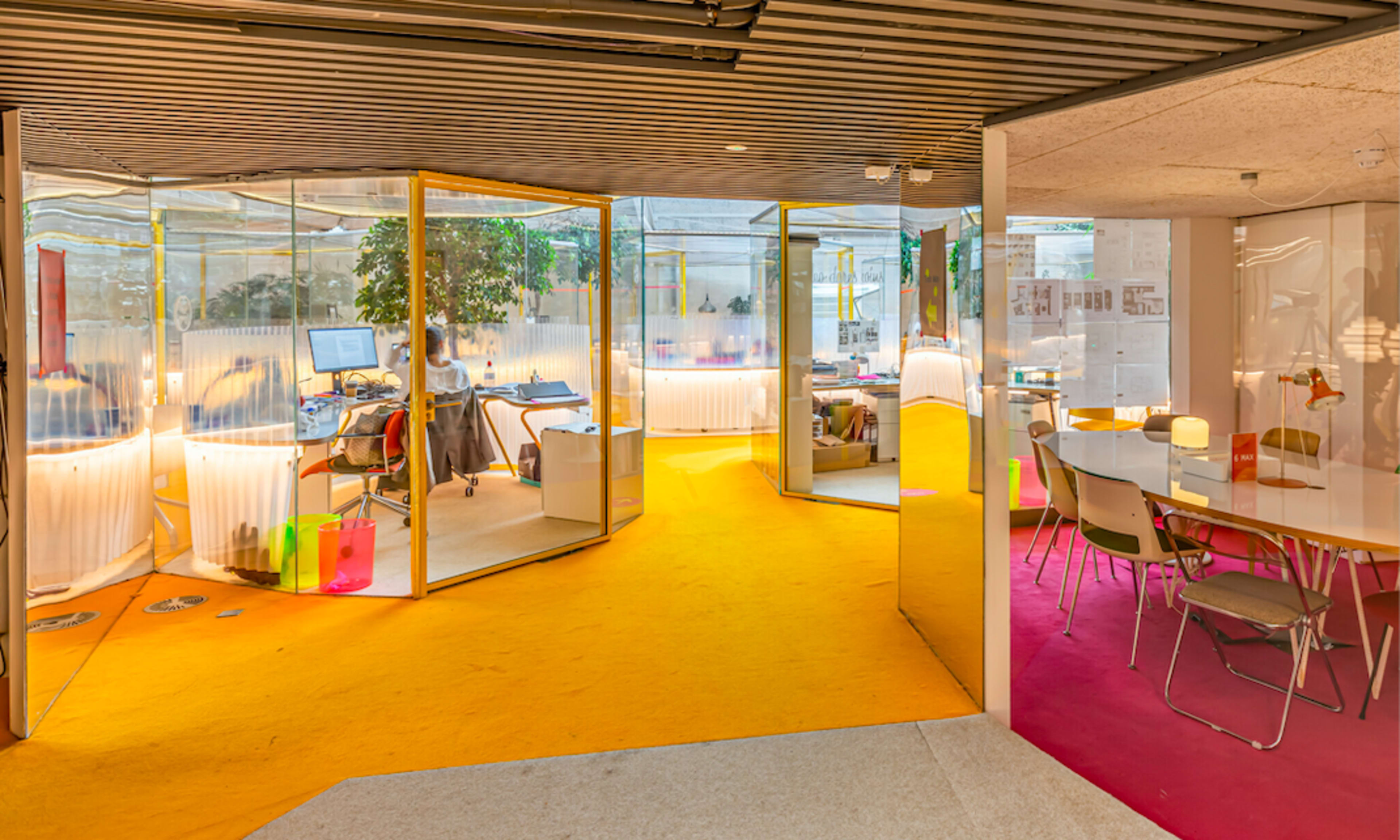 The image shows a modern office space with glass partitions, colorful flooring, and various workstations illuminated by lamps.
