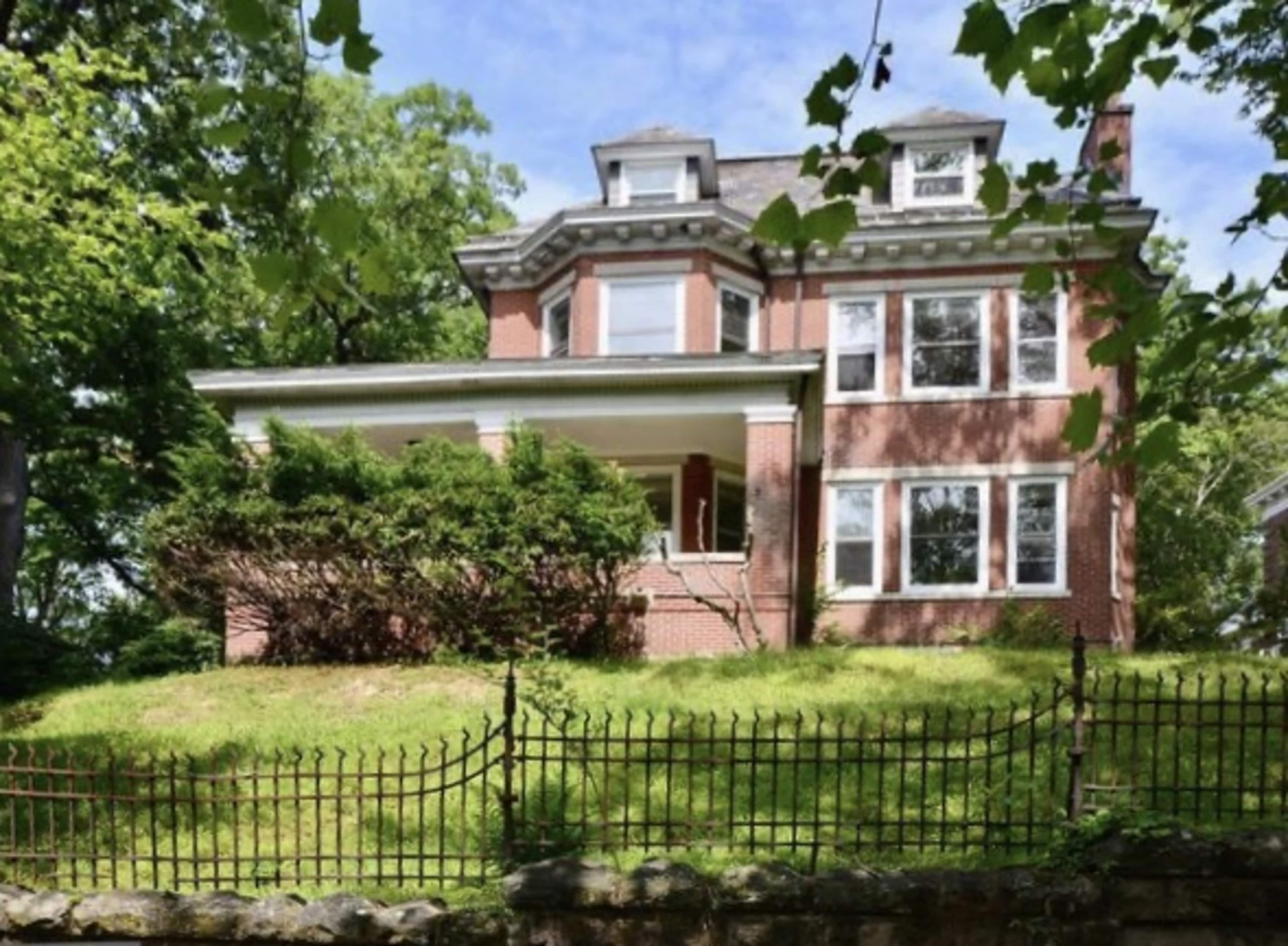 A large, brick Victorian-style house stands on a grassy lot surrounded by a wrought iron fence.