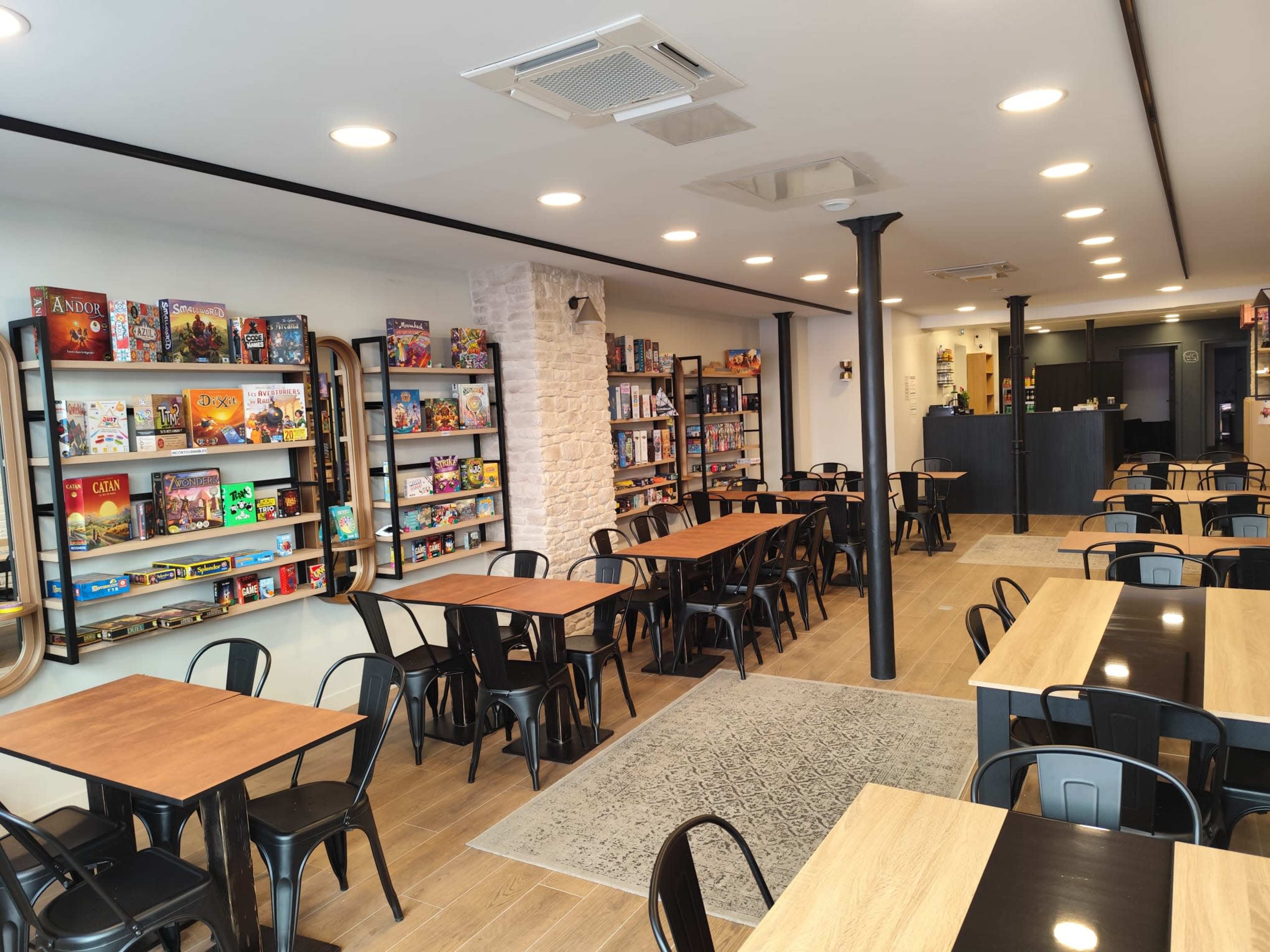 The image shows a game café with rows of tables, shelves filled with board games on the walls, and a counter in the background.