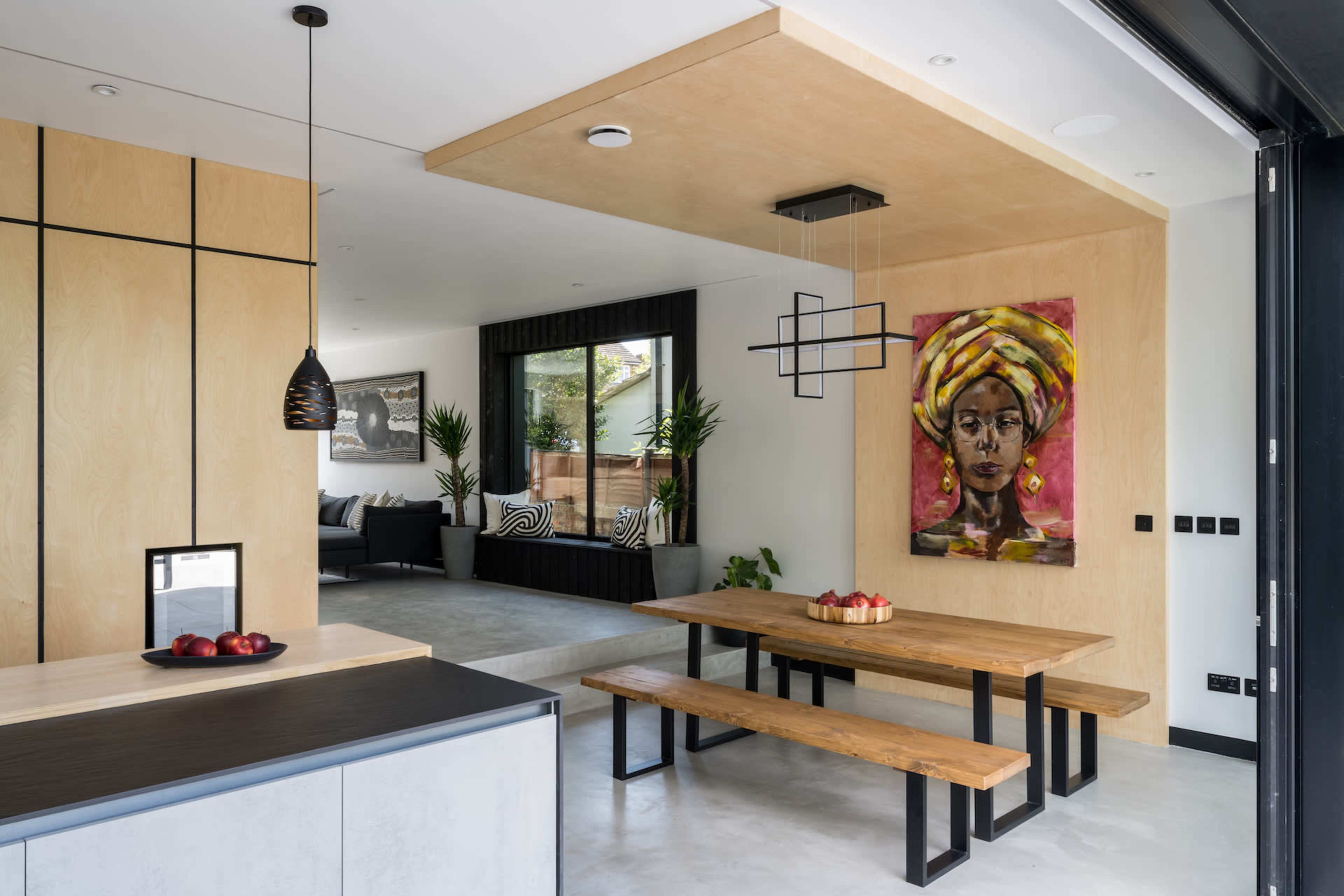 A modern dining area featuring a large wooden table with benches, surrounded by indoor plants and a colorful wall art piece, connecting to a living space.