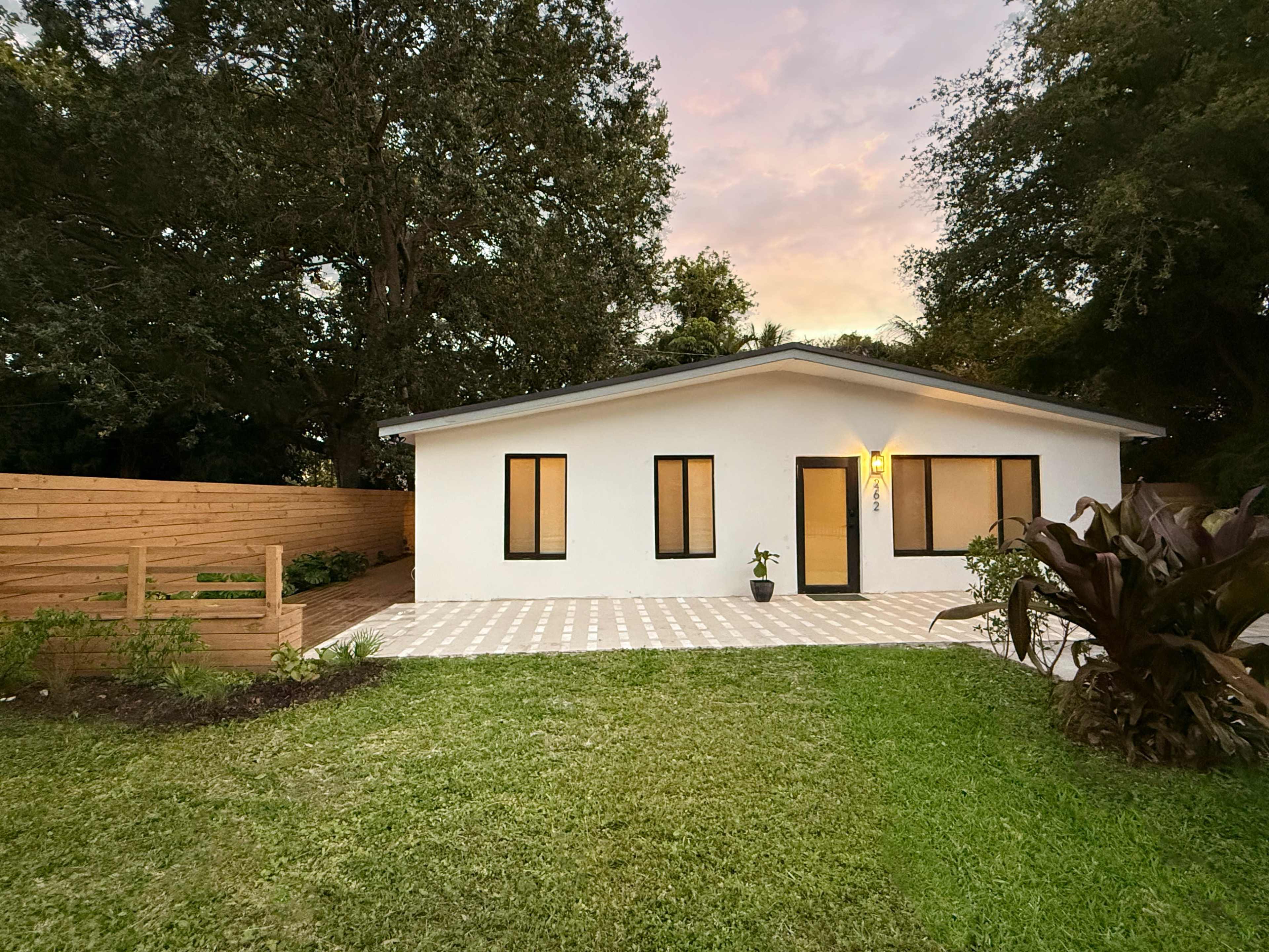 The image shows a modern white house with large windows, surrounded by a grassy lawn and bordered by a wooden fence.