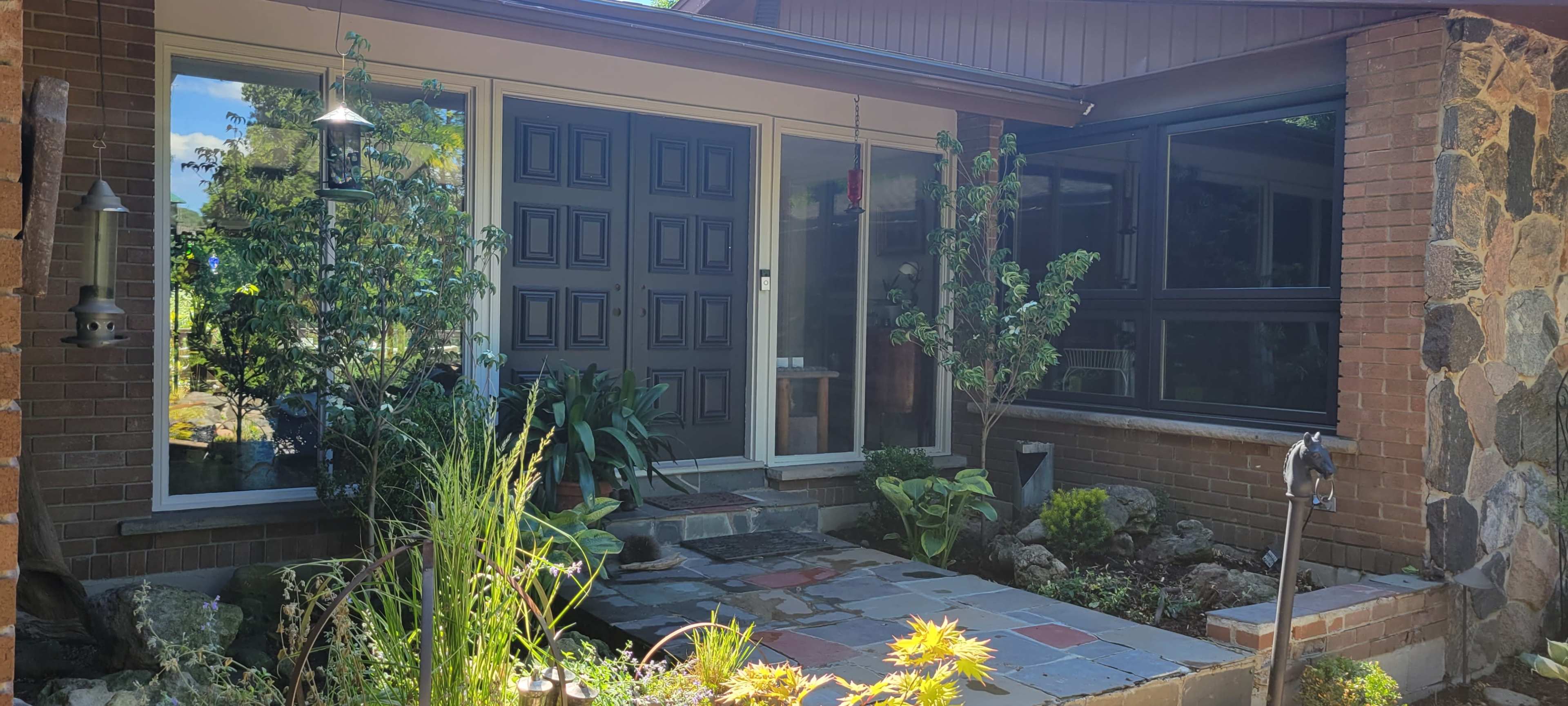 A front entrance of a house featuring double black doors, large windows, and a stone pathway surrounded by plants and landscaping.