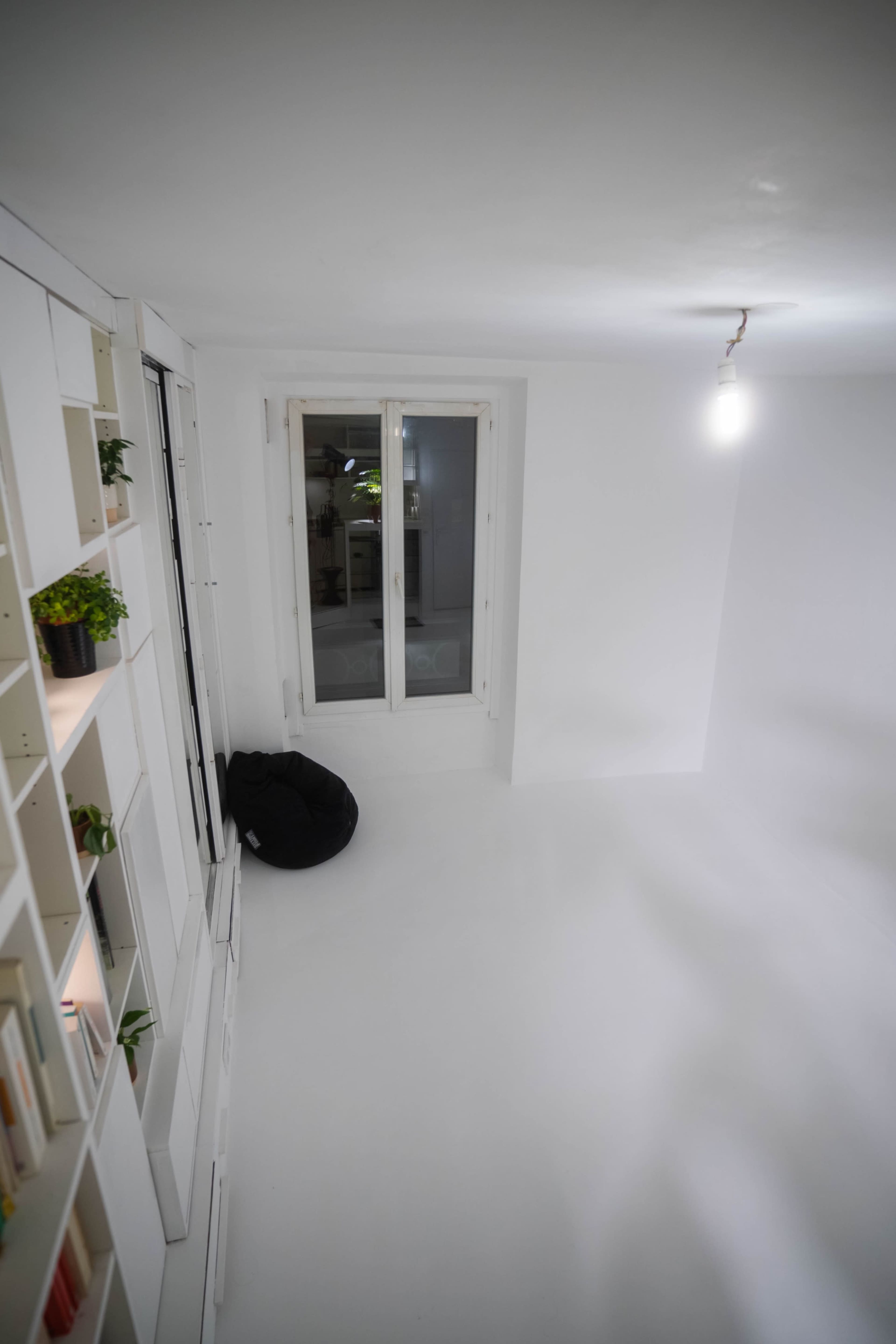 The image shows a minimalist white room with a bookcase, a bean bag chair in the corner, and a window letting in natural light.