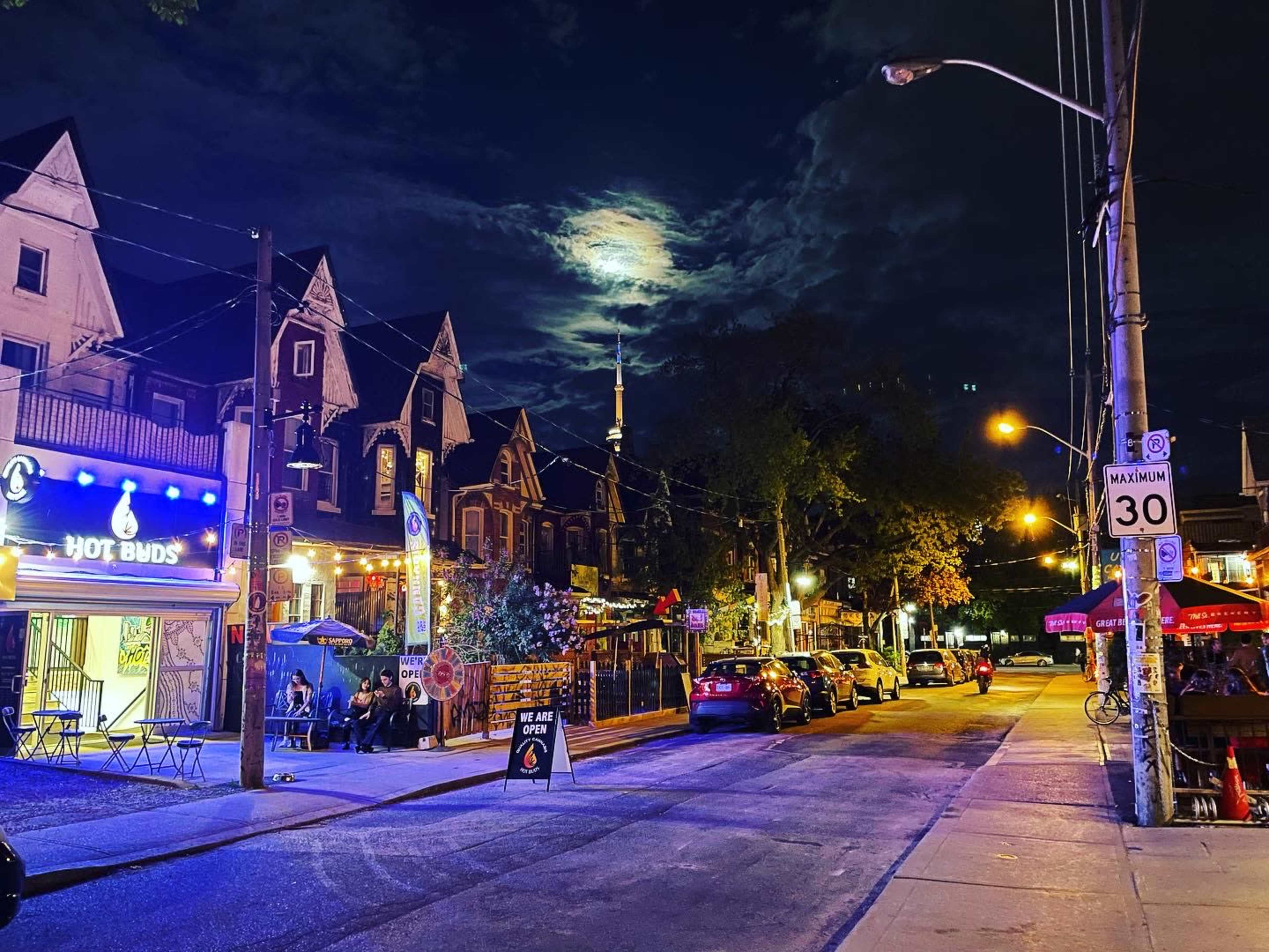 A quiet street at night features illuminated storefronts, parked cars, and a large moon partially obscured by clouds.