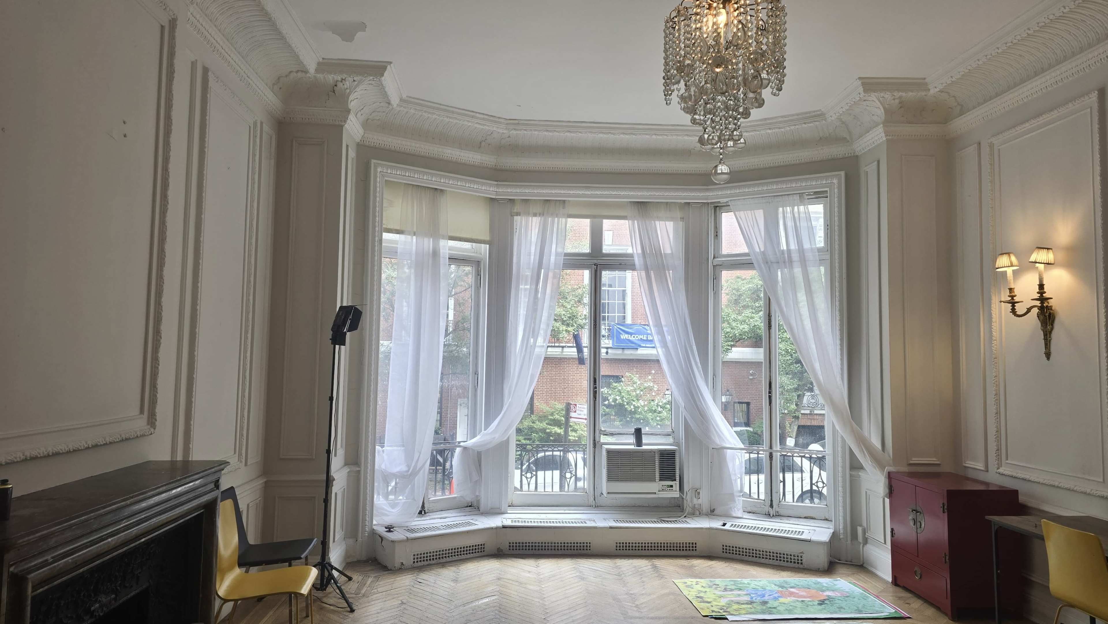 The image shows a spacious room with large bay windows, a chandelier, and a mix of furniture, including a yellow chair and a red cabinet.