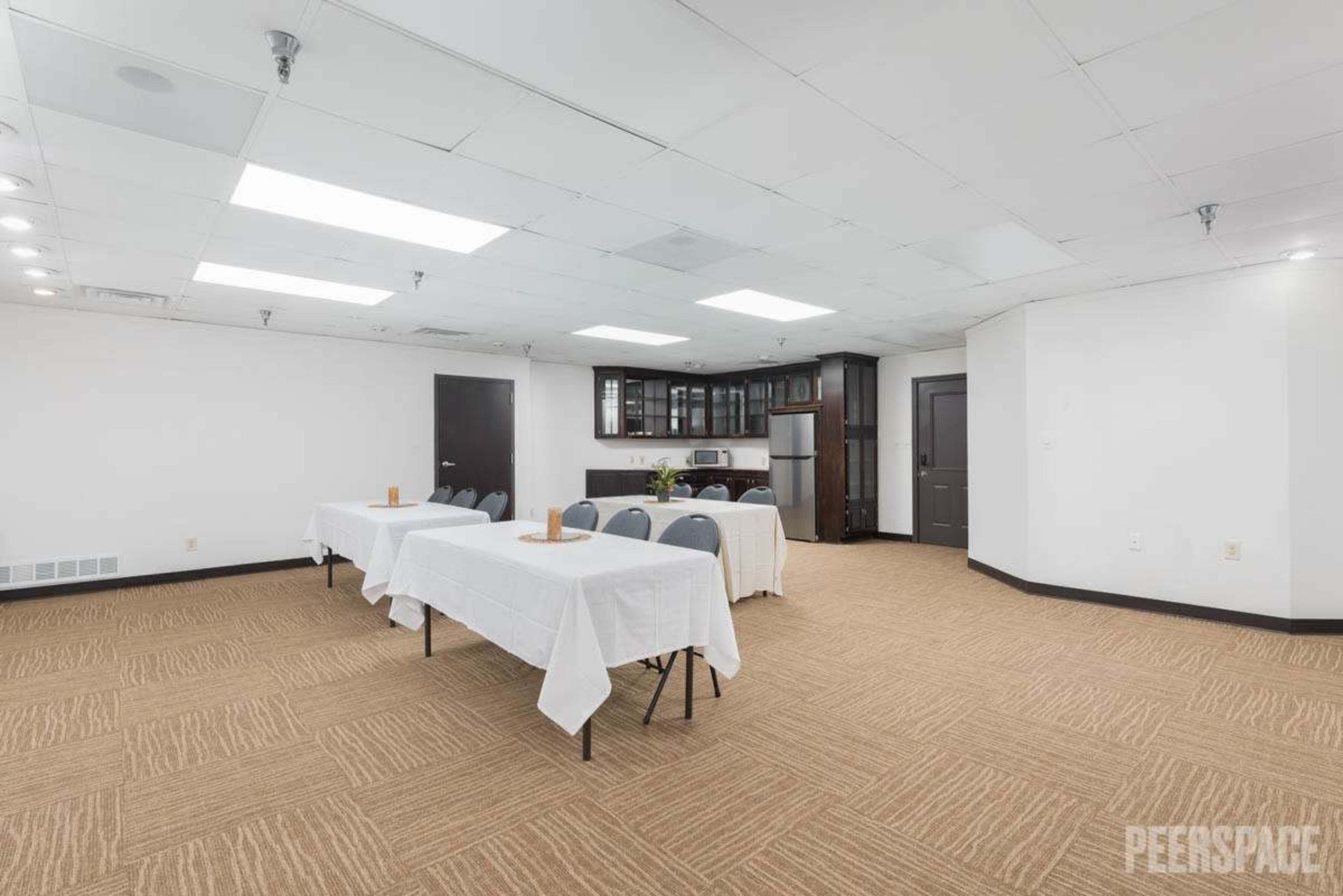 The image shows a meeting room with two long tables covered in white tablecloths and arranged with chairs, along with a kitchenette area in the background.