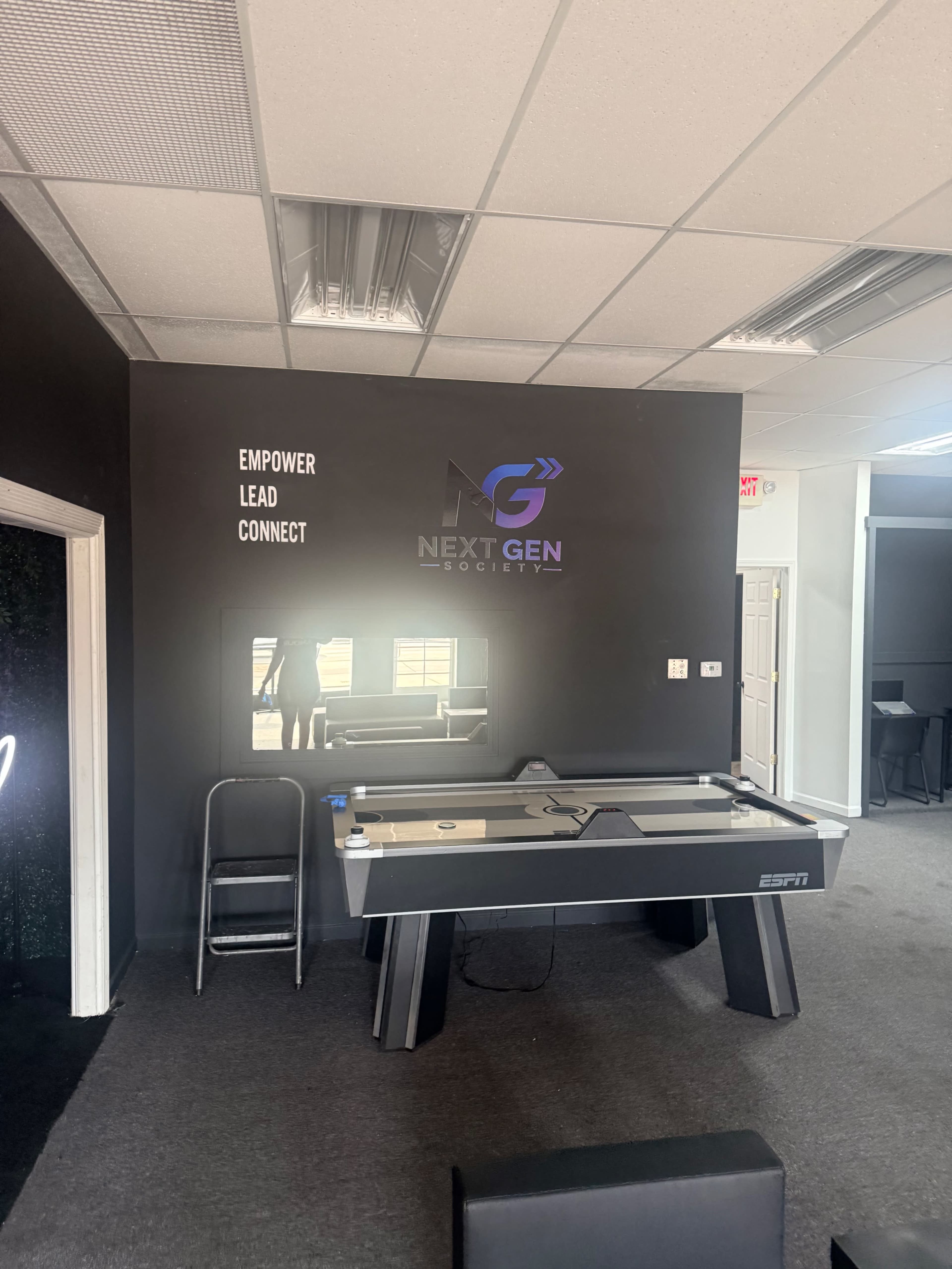 The image shows a room with a black wall featuring the text "EMPOWER LEAD CONNECT," a logo for "NEXT GEN SOCIETY," and an air hockey table beside a folding chair.