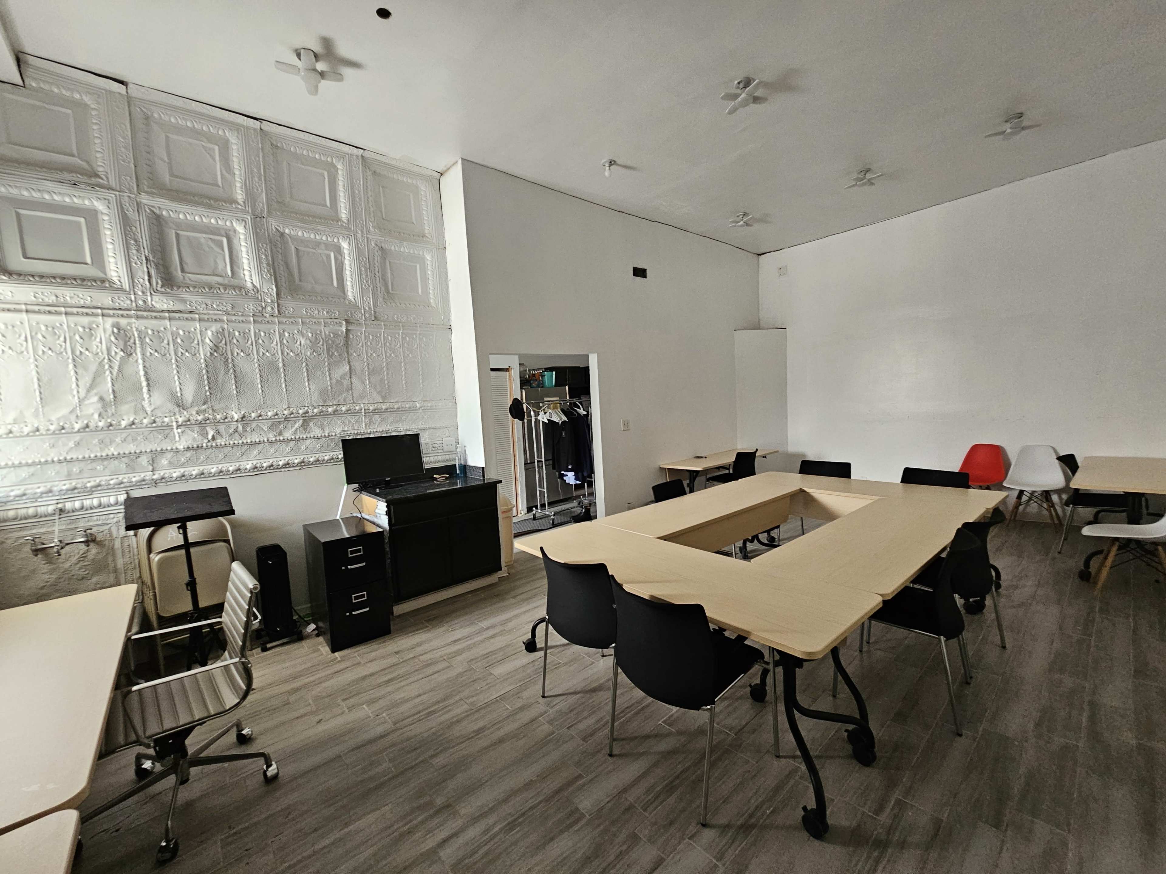 The image shows a bright, spacious room with multiple tables and chairs arranged for work or meetings, featuring a textured wall and simple furnishings.