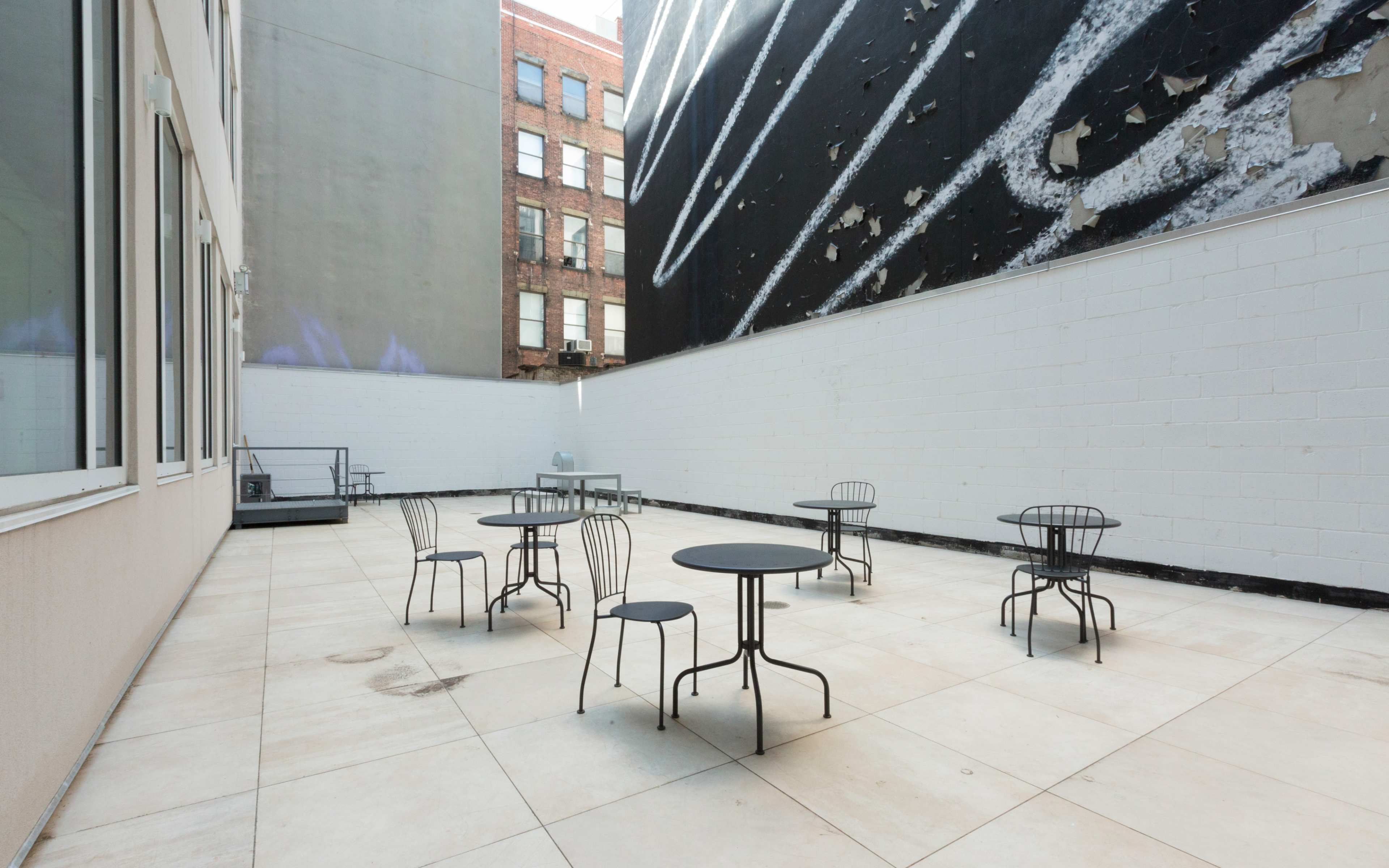 An outdoor terrace with several black metal tables and chairs on a tiled surface, adjacent to a wall featuring a large mural.