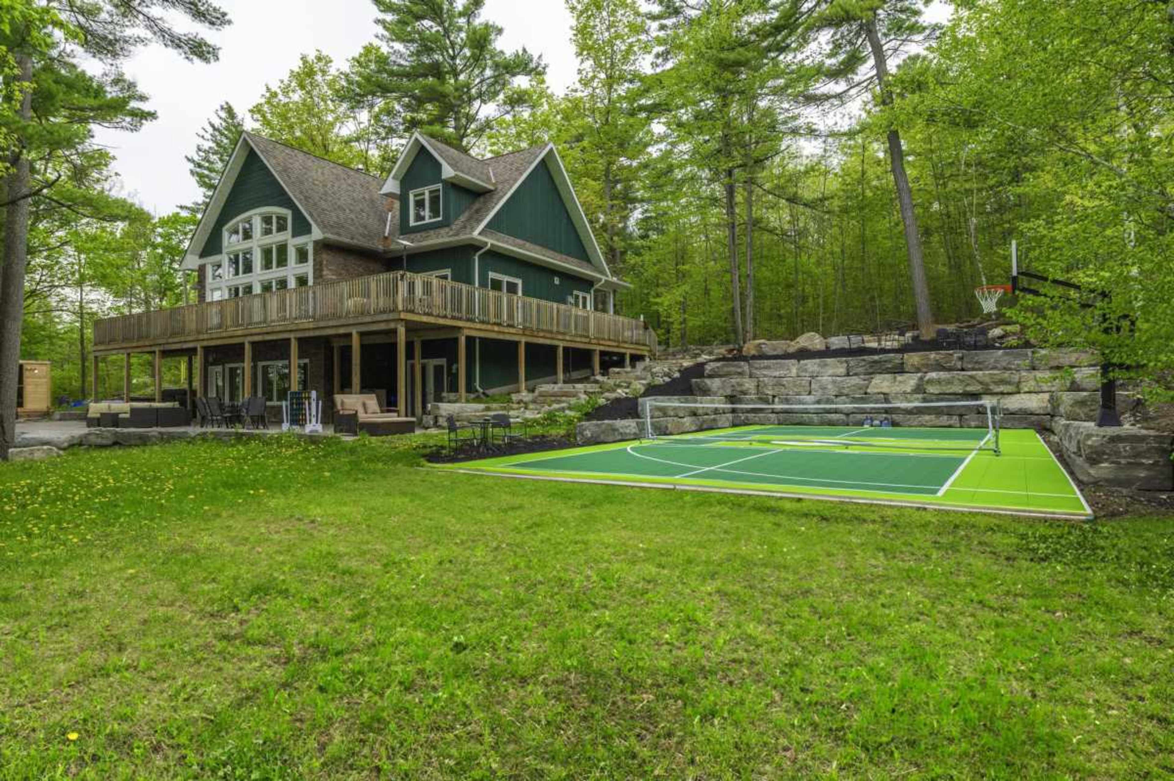 A large green and brown house with a deck overlooks a grass yard with a sports court and surrounding trees.