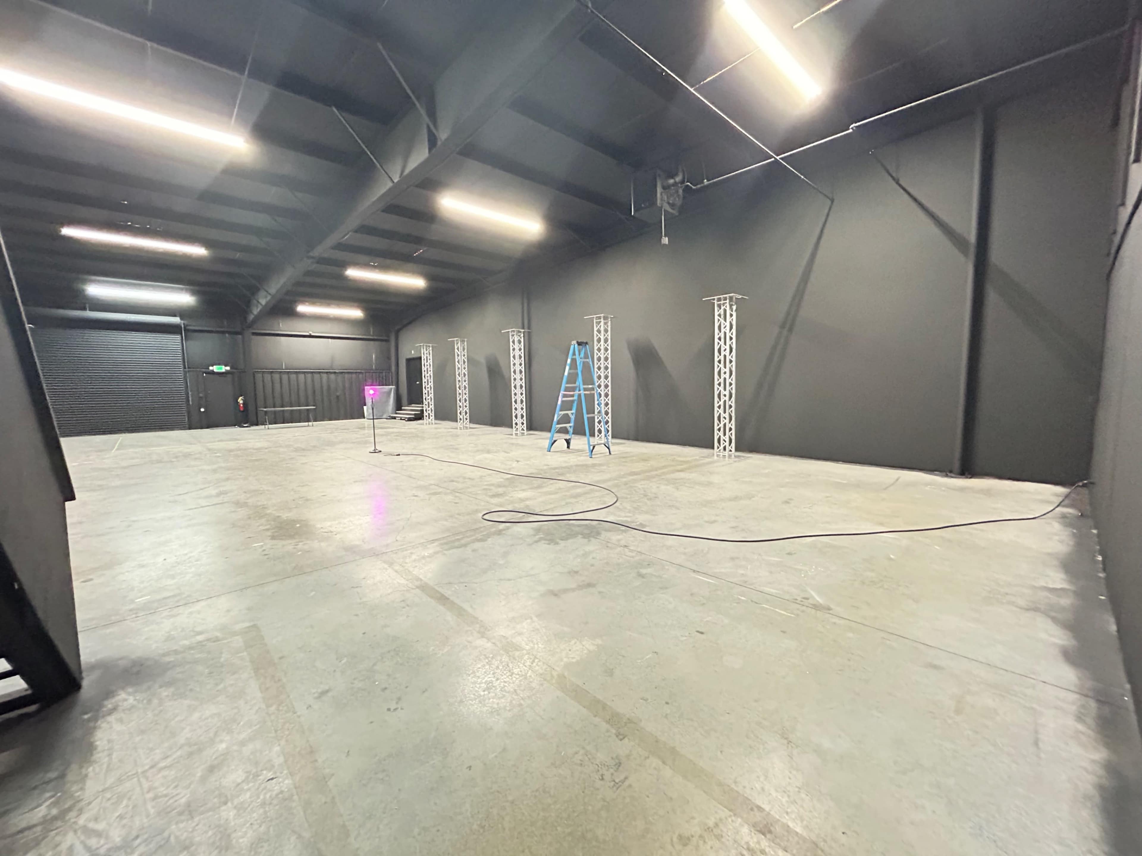 A spacious, empty interior of a warehouse or studio, featuring black walls, bare concrete floor, and several light trusses positioned around the room.