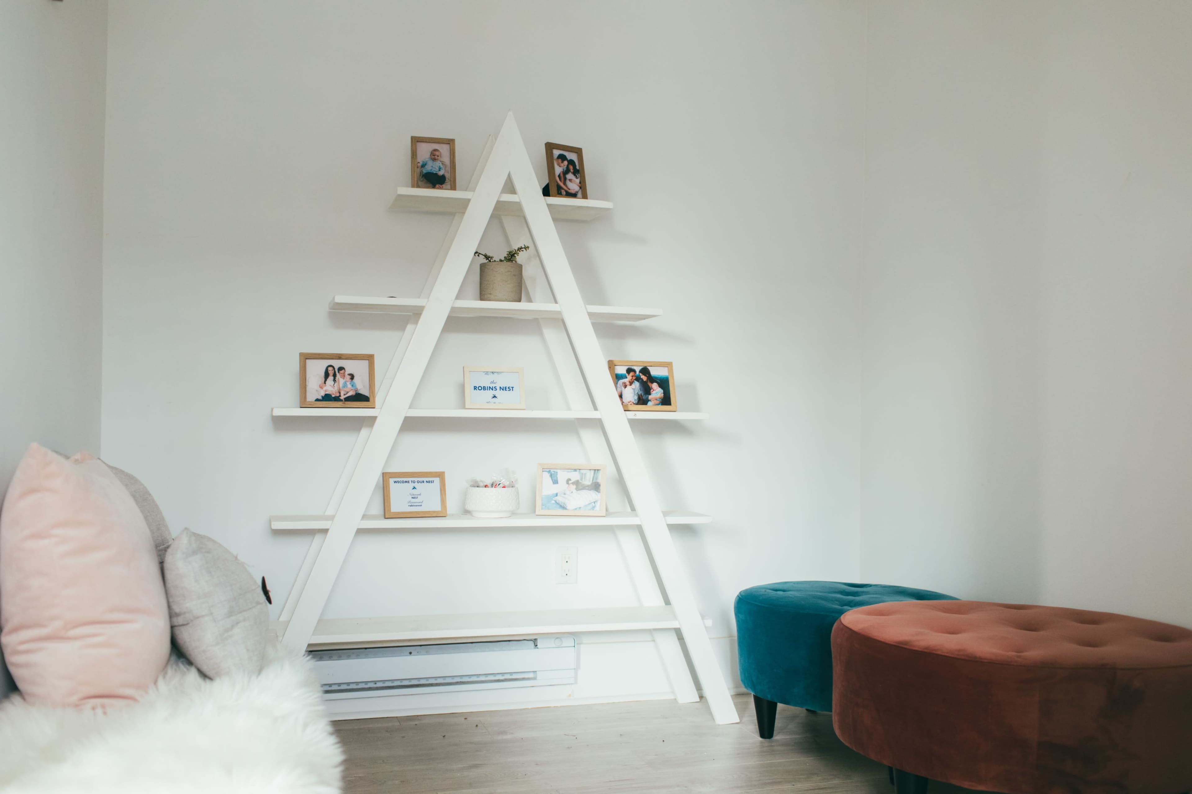 A triangular white bookshelf displays framed photos and a small potted plant against a light-colored wall, with two upholstered ottomans nearby.