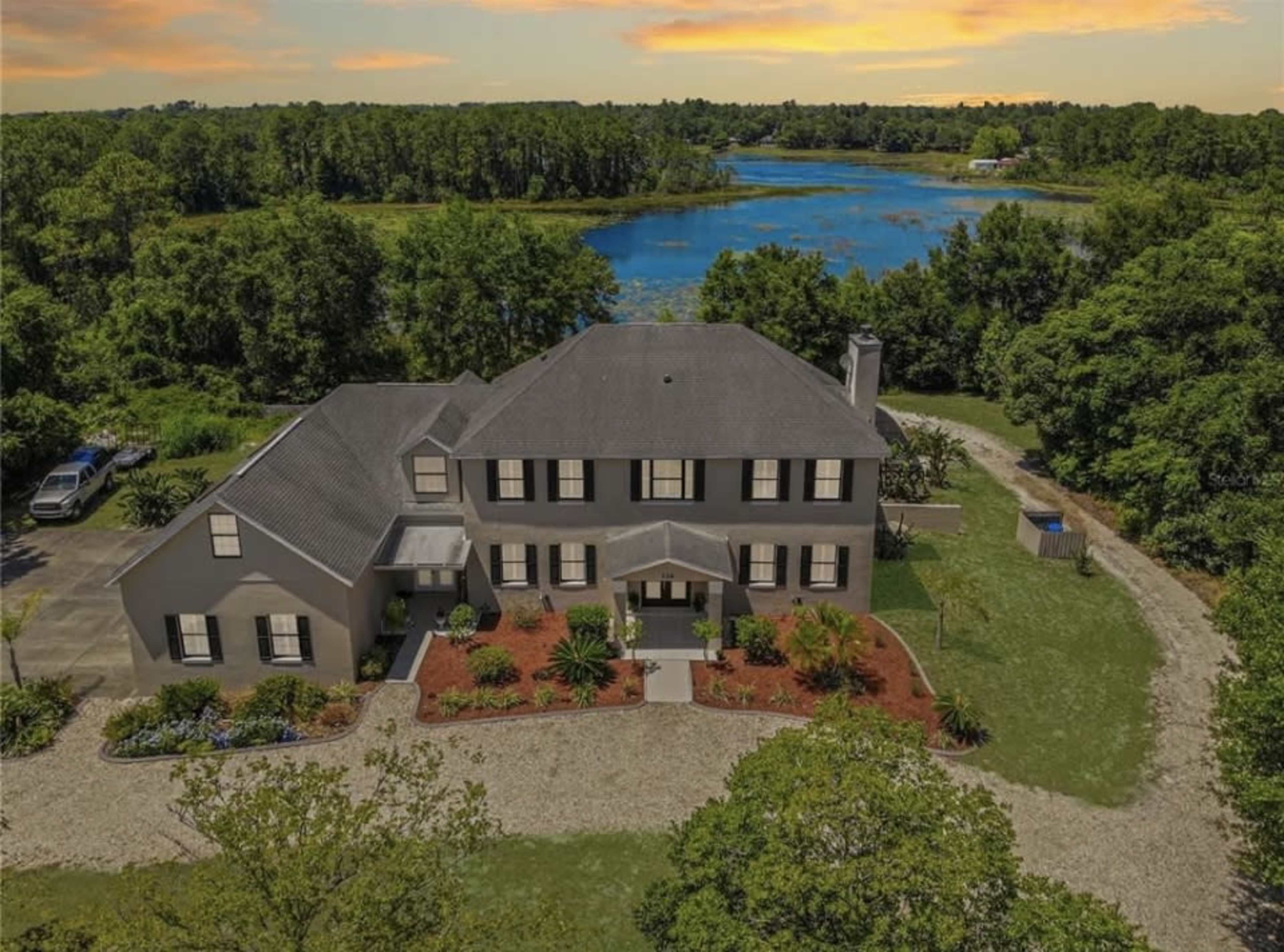 A large two-story house with a dark roof is set on a grassy landscape near a calm body of water, surrounded by trees and landscaping.