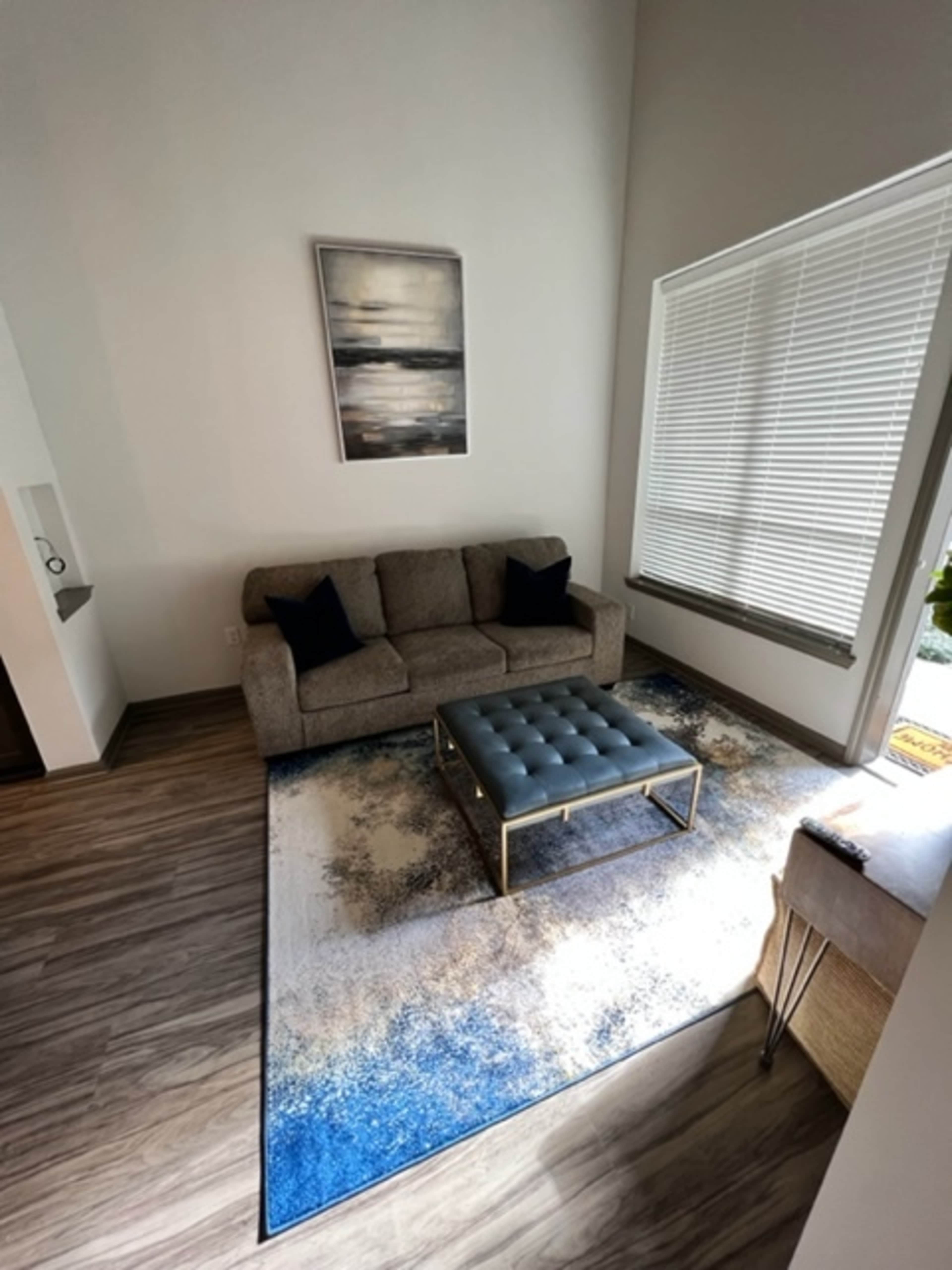 A modern living room features a gray sofa, a blue tufted coffee table, and a large area rug with a blue and beige design under a corner window with blinds.