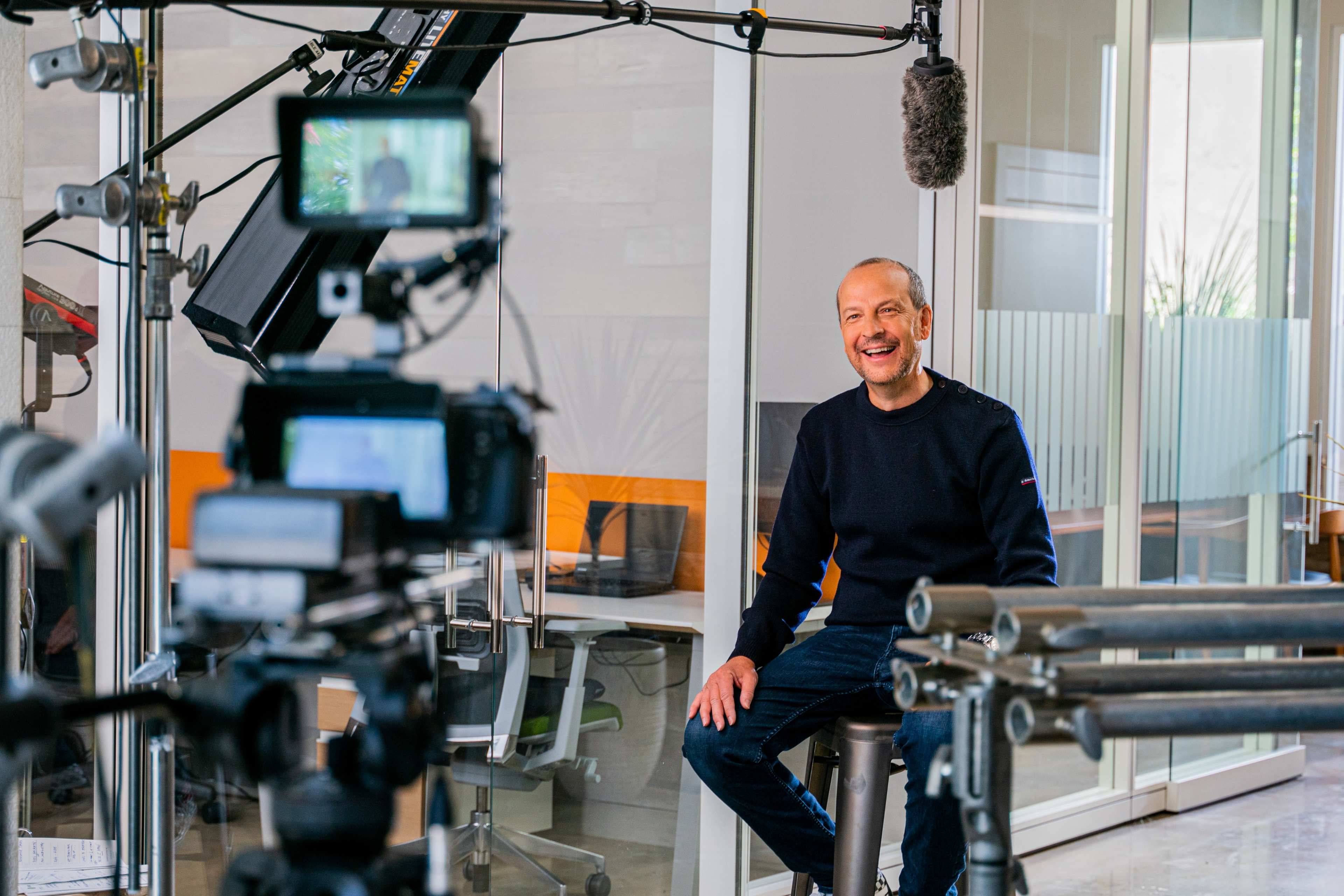 A man sits on a stool in a well-lit studio, preparing for an interview with cameras and microphones set up in front of him.