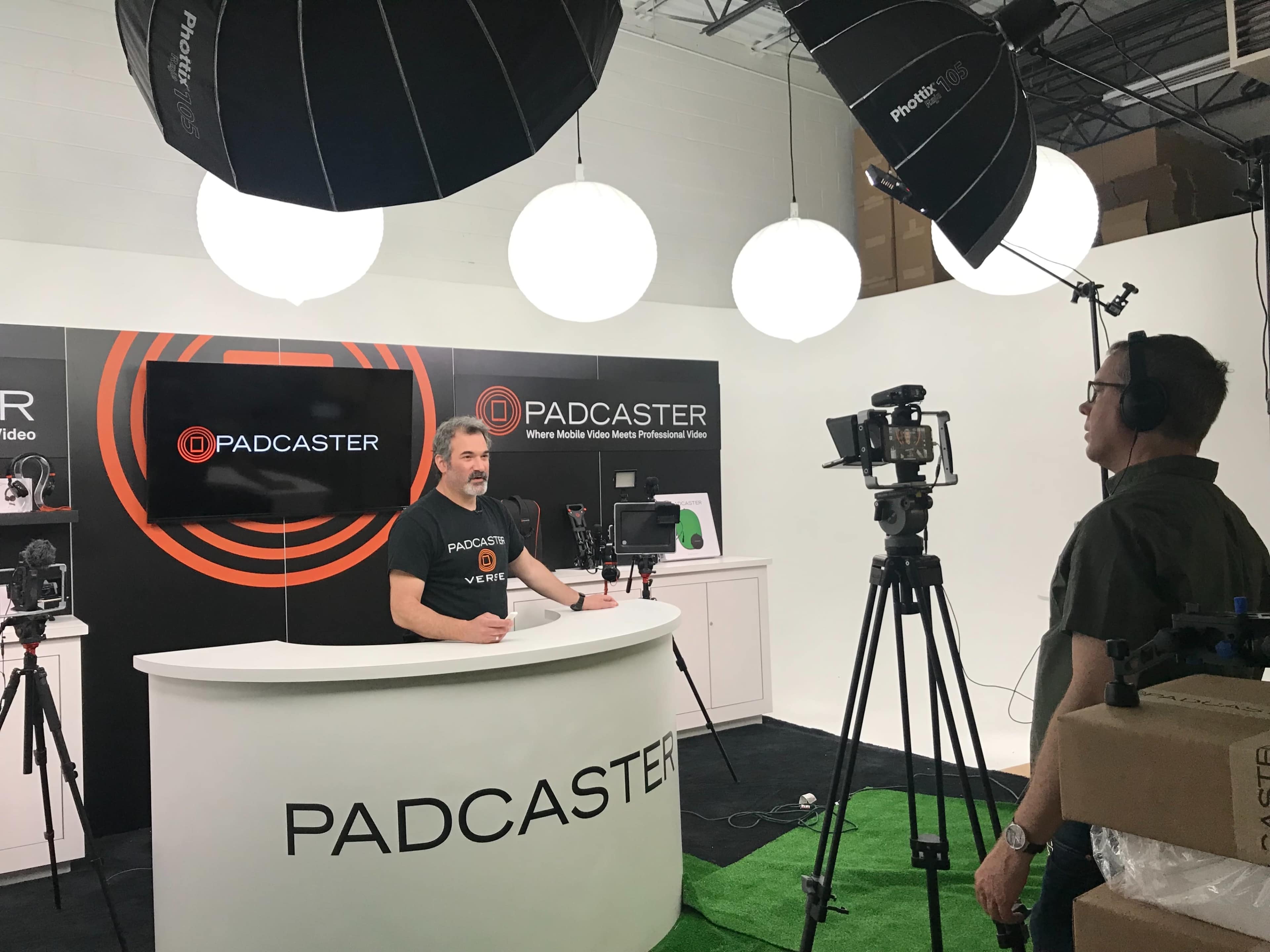 A man is standing behind a curved white table with the logo "PADCASTER" while another man operates a camera under studio lighting.