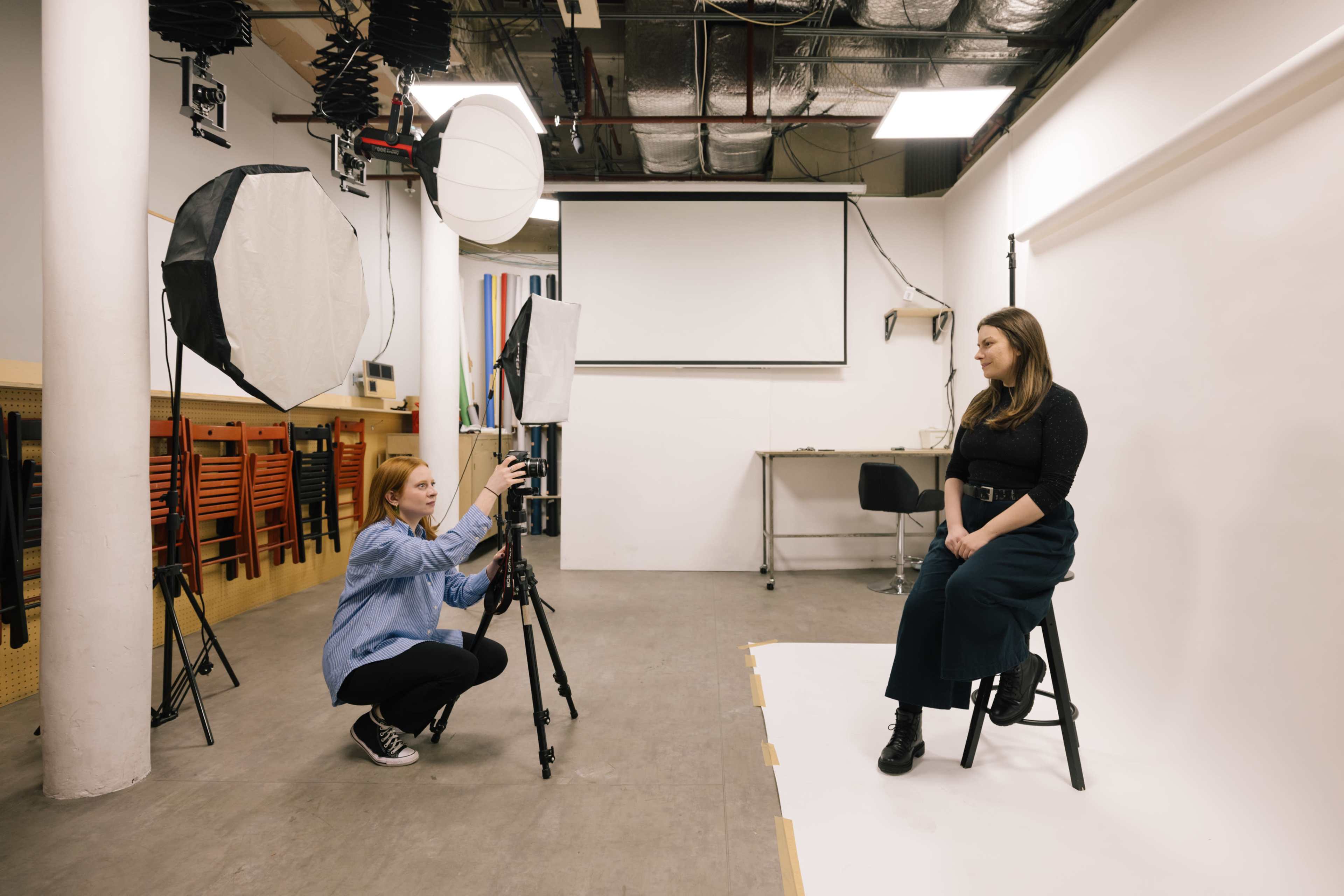 A photographer adjusts equipment while a model poses on a stool in a studio setting.