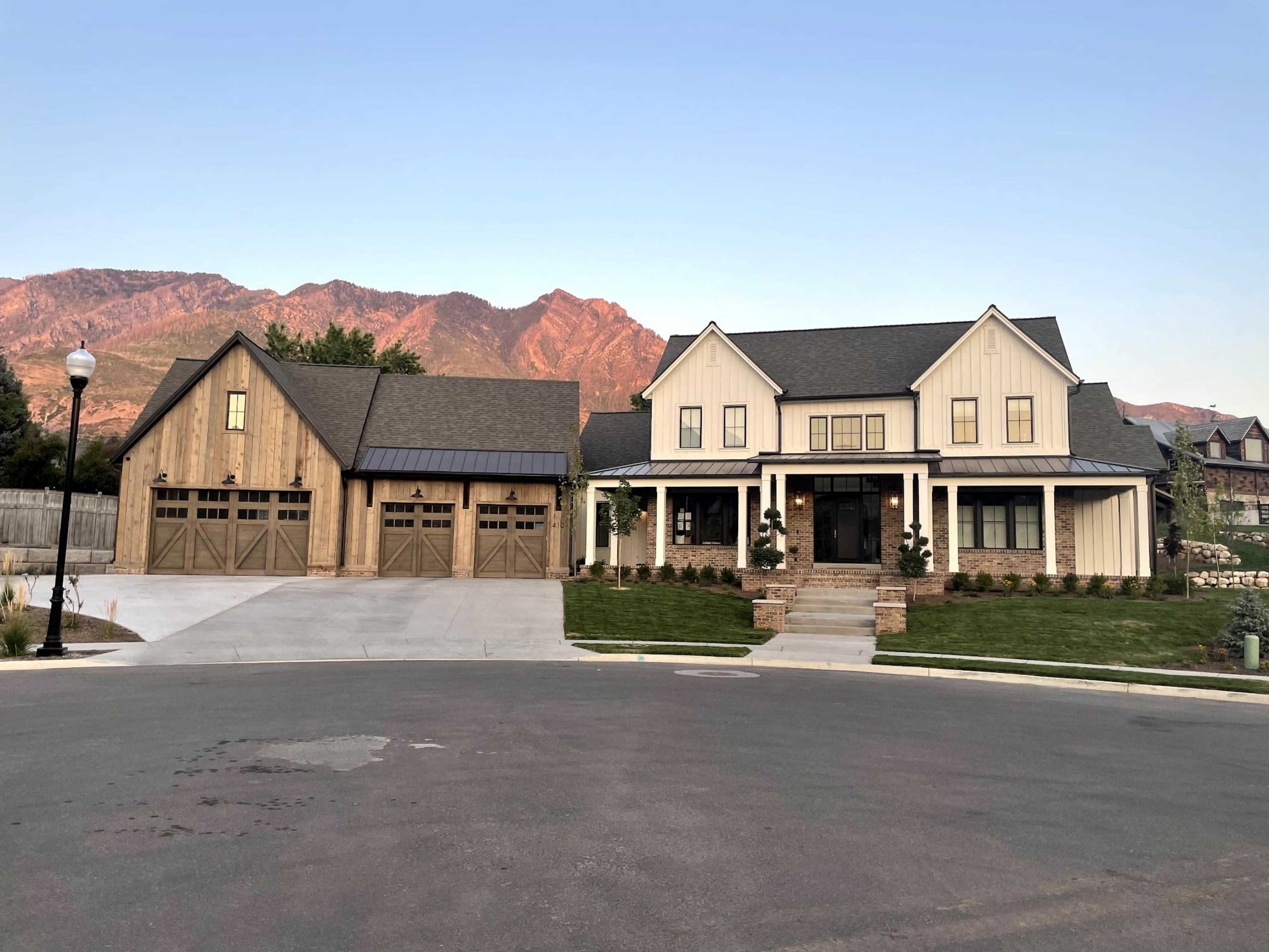 The image shows two large homes with a landscaped yard and a mountain backdrop, situated on a curved street.
