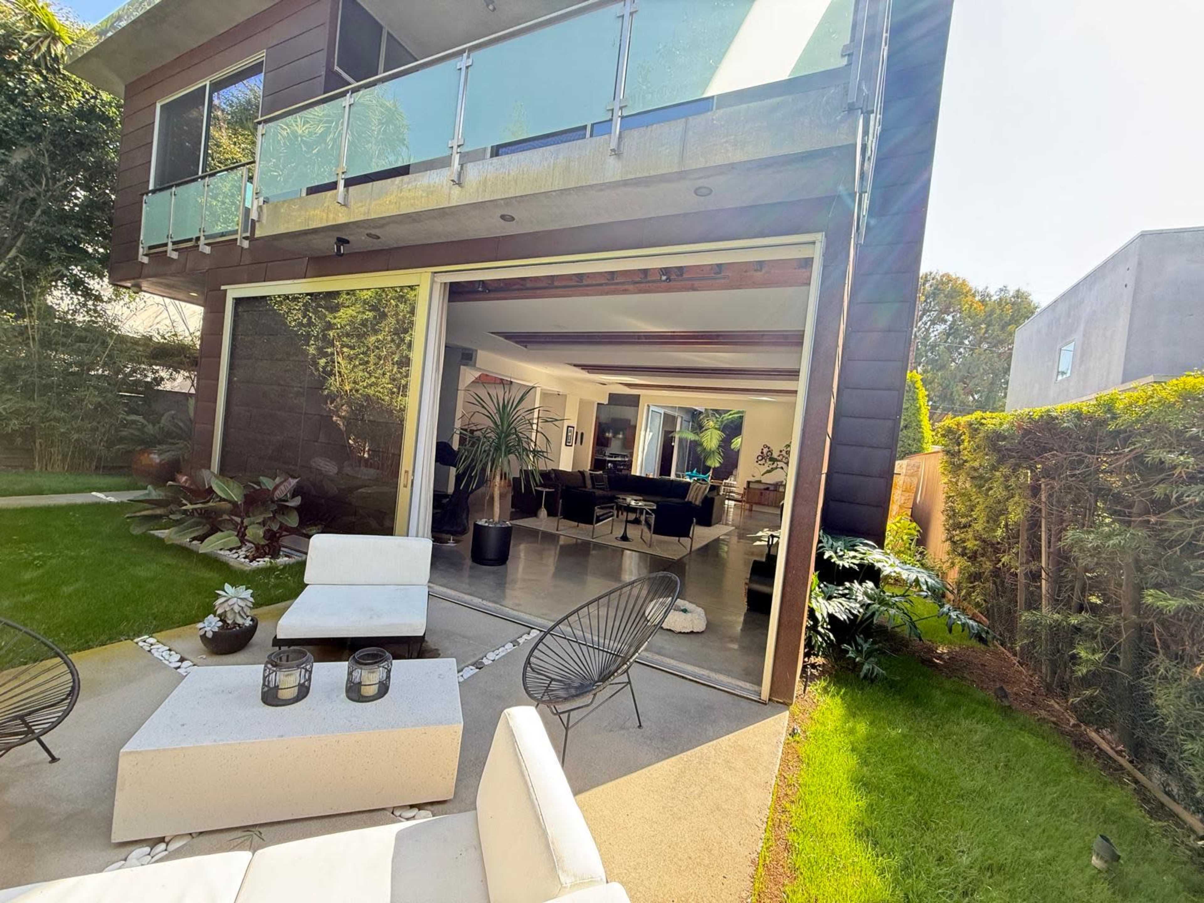 A modern house features large glass doors opening to a patio with outdoor seating and a well-maintained garden.