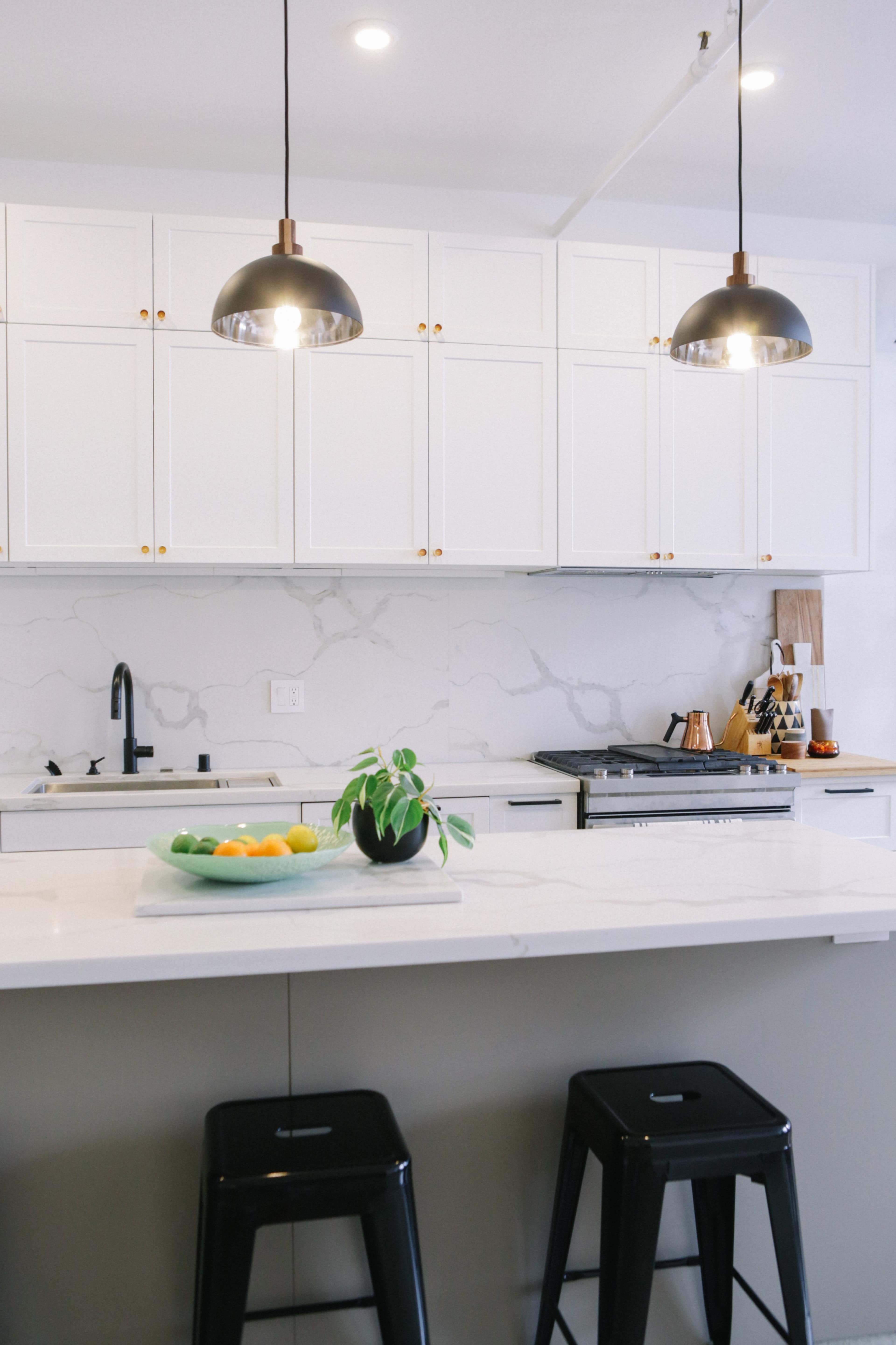 A modern kitchen features white cabinets, a marble backsplash, and two pendant lights above a countertop with bar stools.