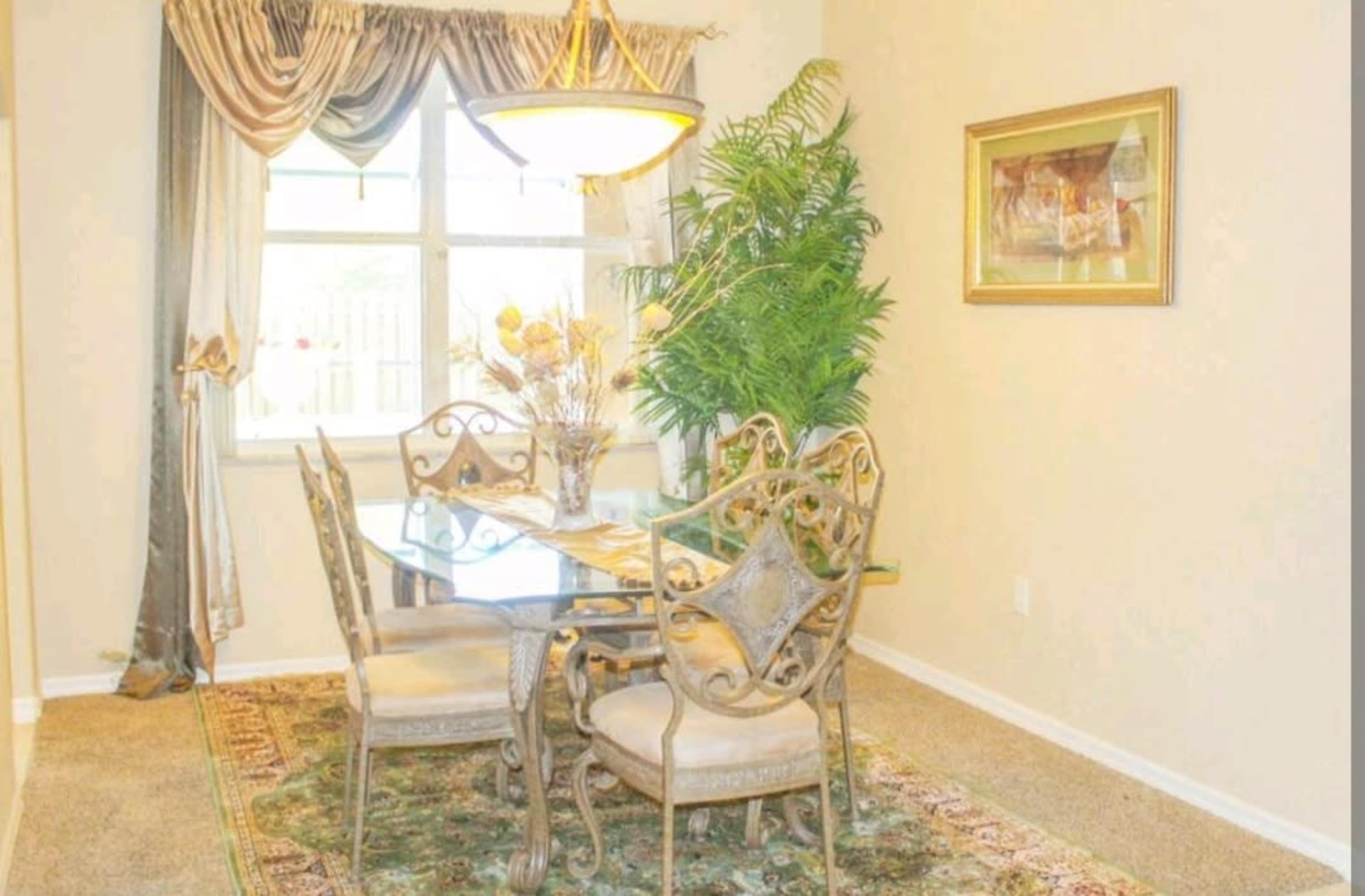 A dining area features a glass table surrounded by four ornate chairs, with a decorative plant and a painting accentuating the space.