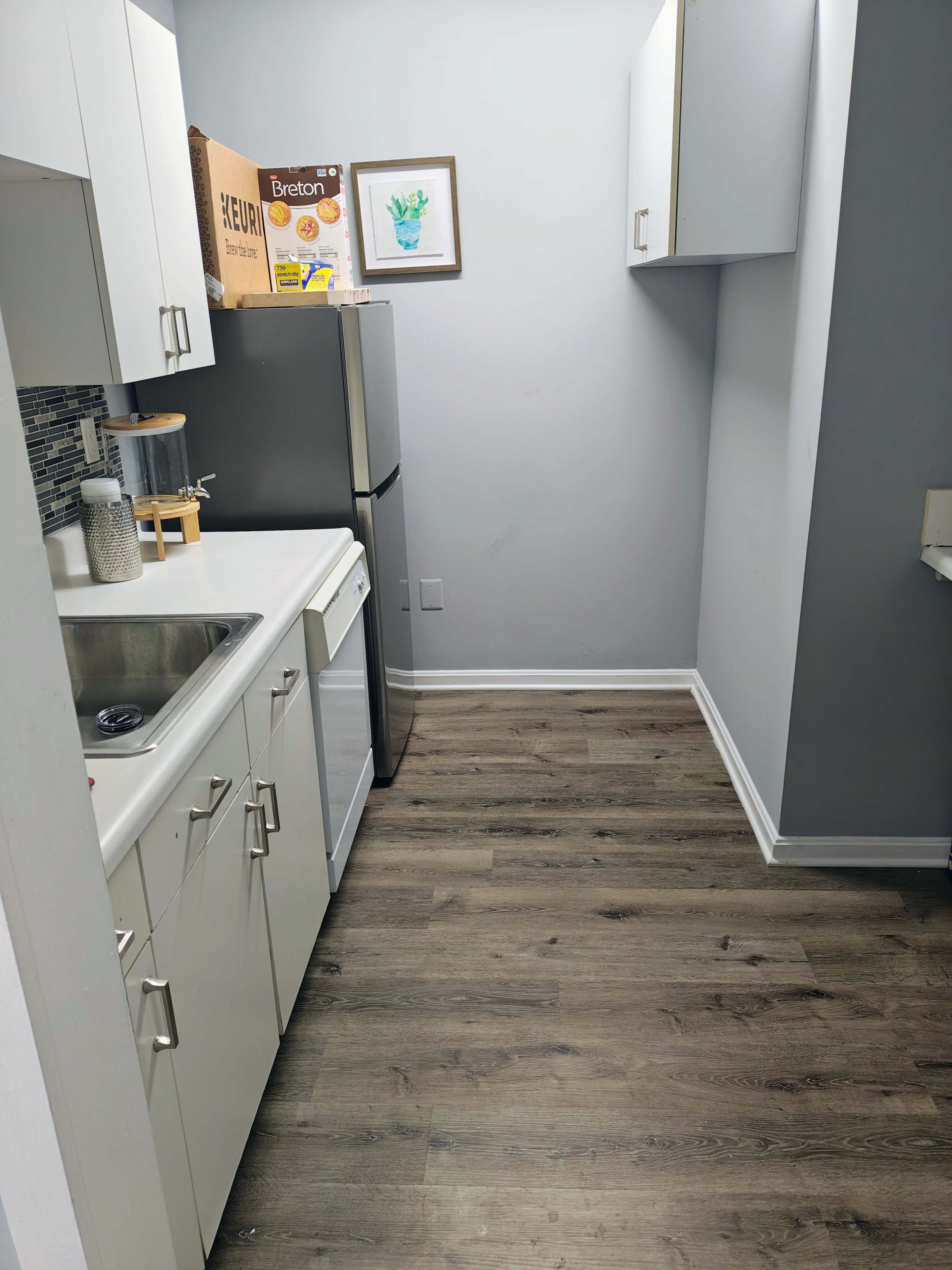 The image shows a narrow kitchen with white cabinetry, a silver refrigerator, and a single sink with a wooden countertop.