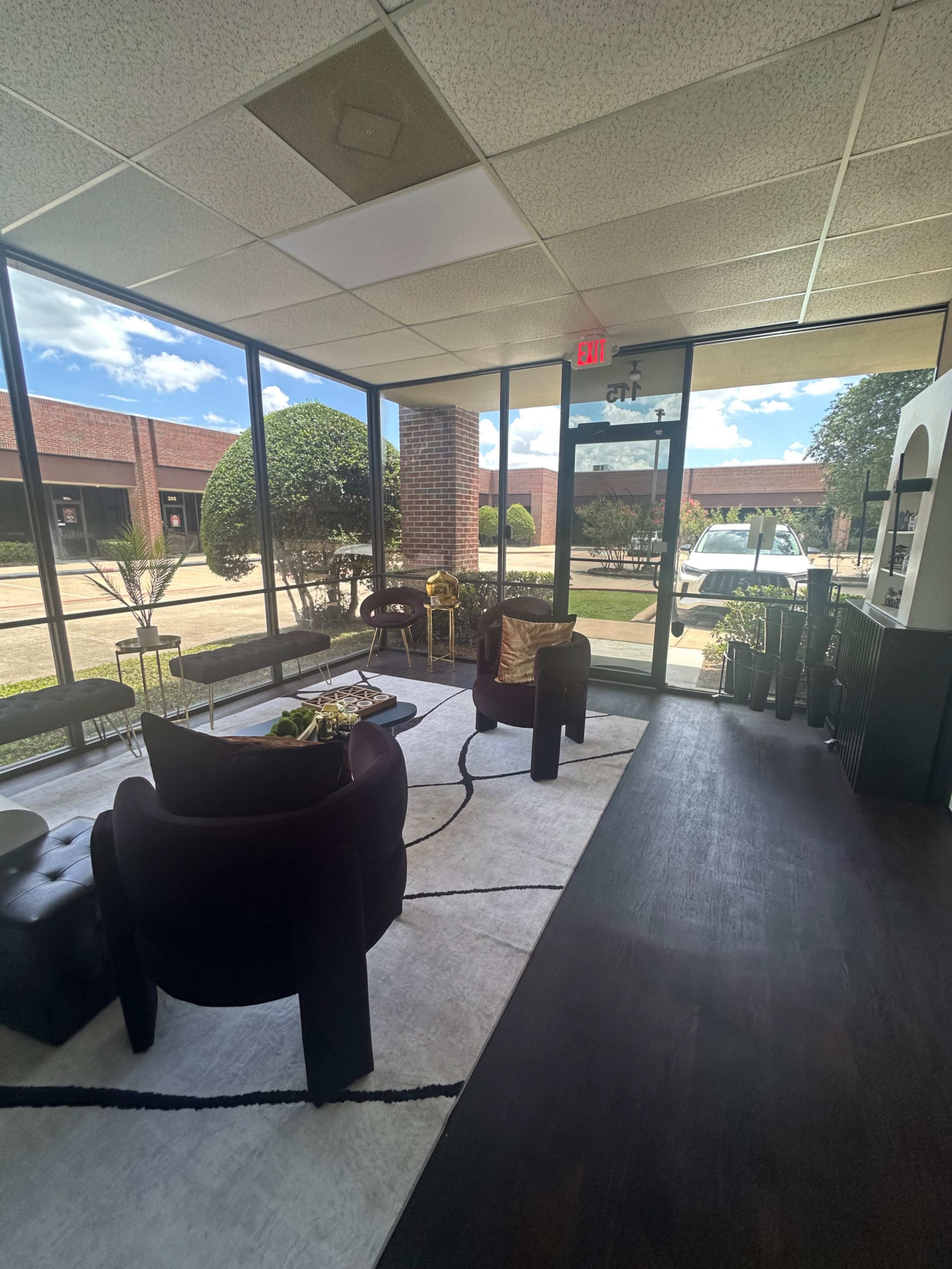 The image shows a modern waiting area with large windows, featuring two chairs, a coffee table, and a decorative rug.