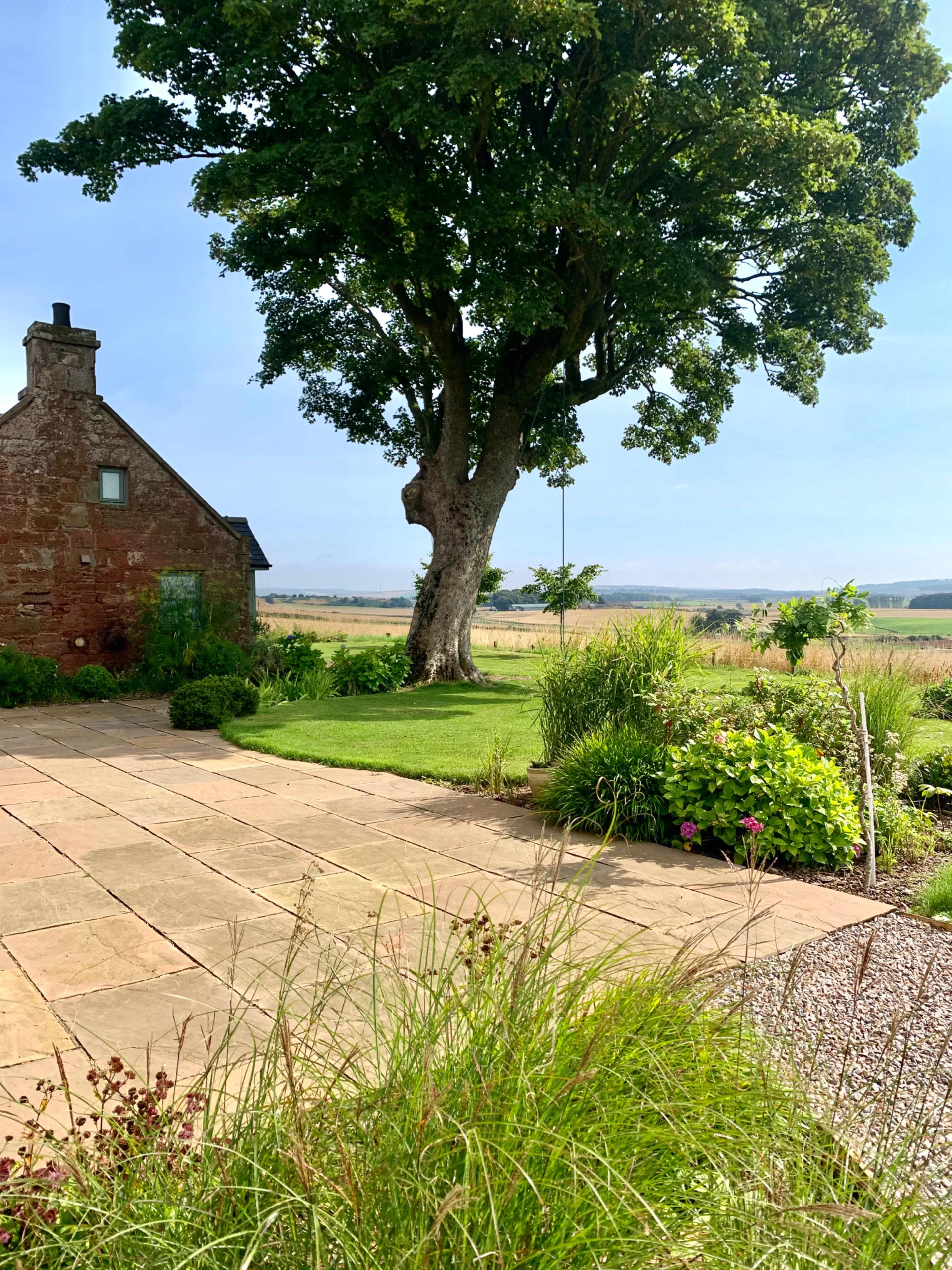 A stone cottage stands beside a large tree, with a stone patio and landscaped garden overlooking a grassy field.