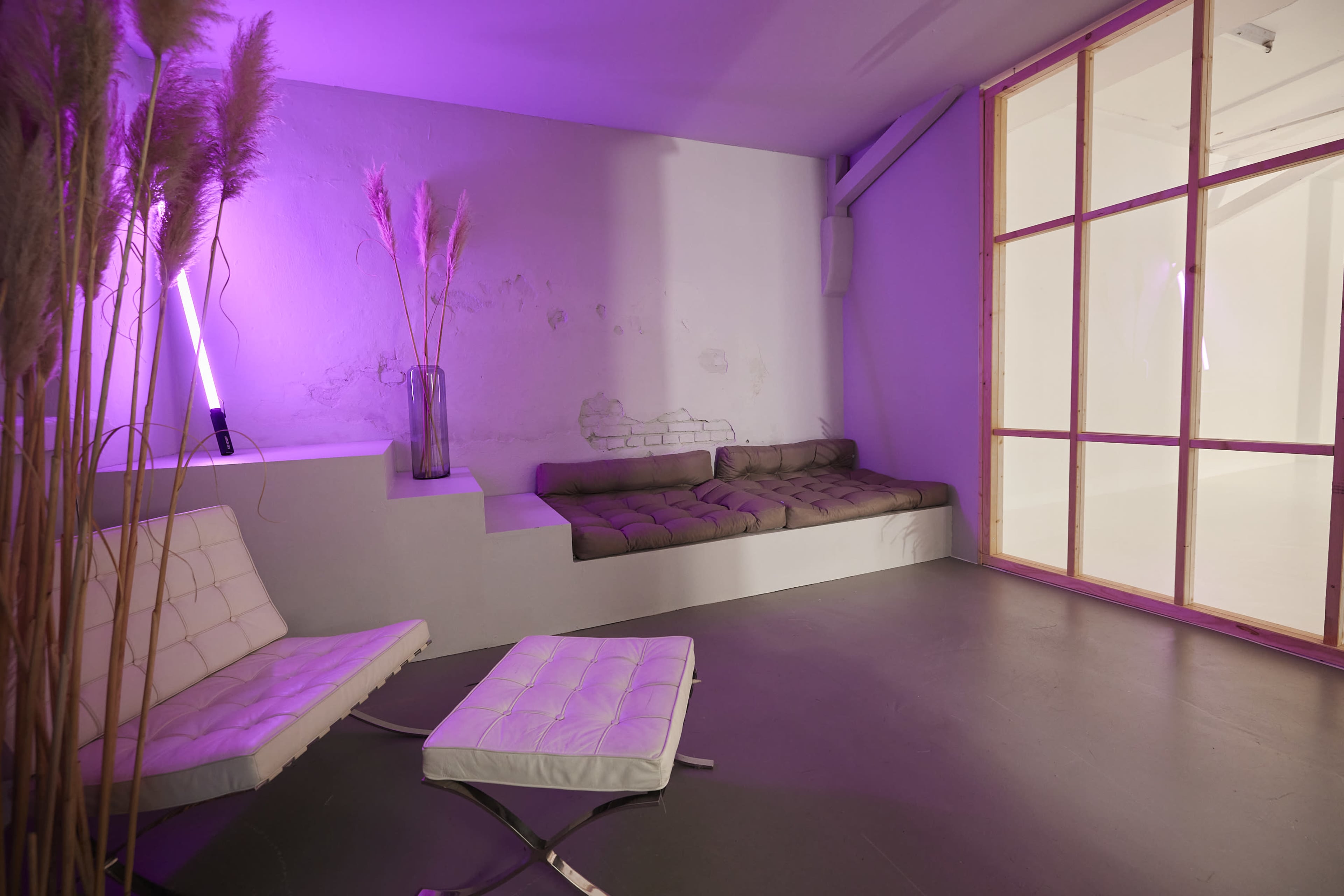 The image shows a minimalist interior space with a low seating area and a glass partition, illuminated by purple light.