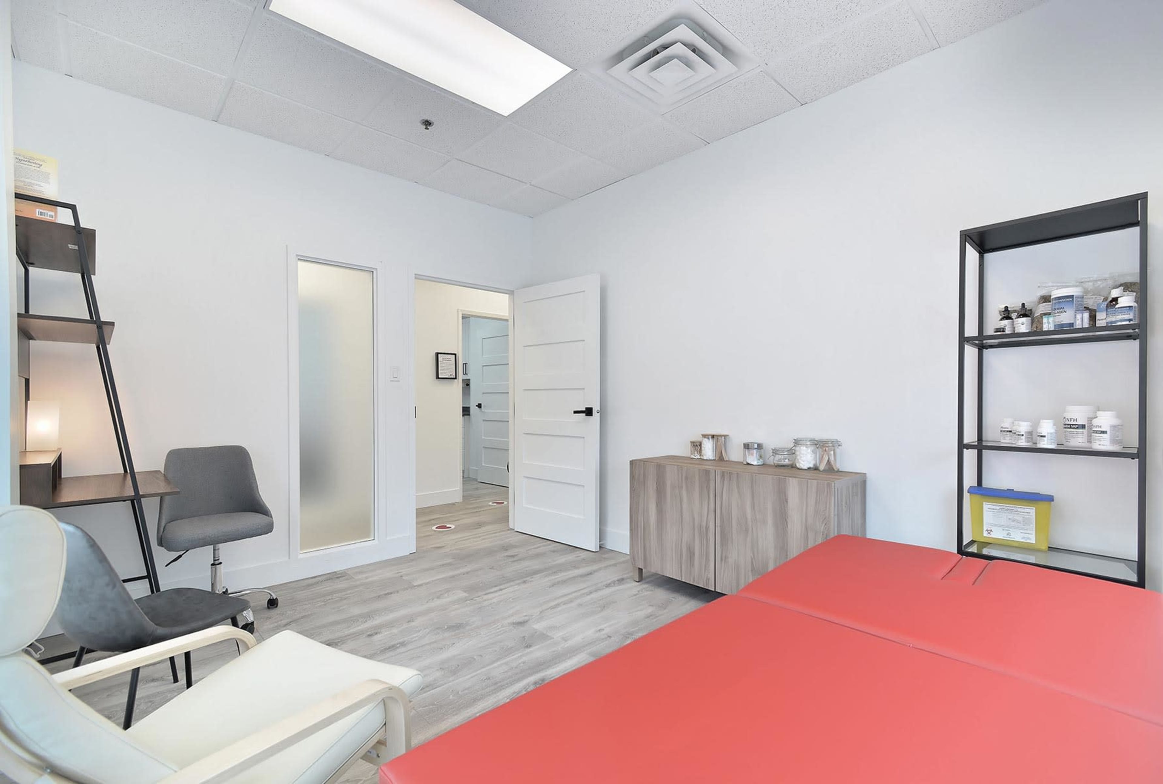 A minimalist therapy room featuring a red treatment table, a gray chair, a shelving unit with containers, and a door leading to another area.