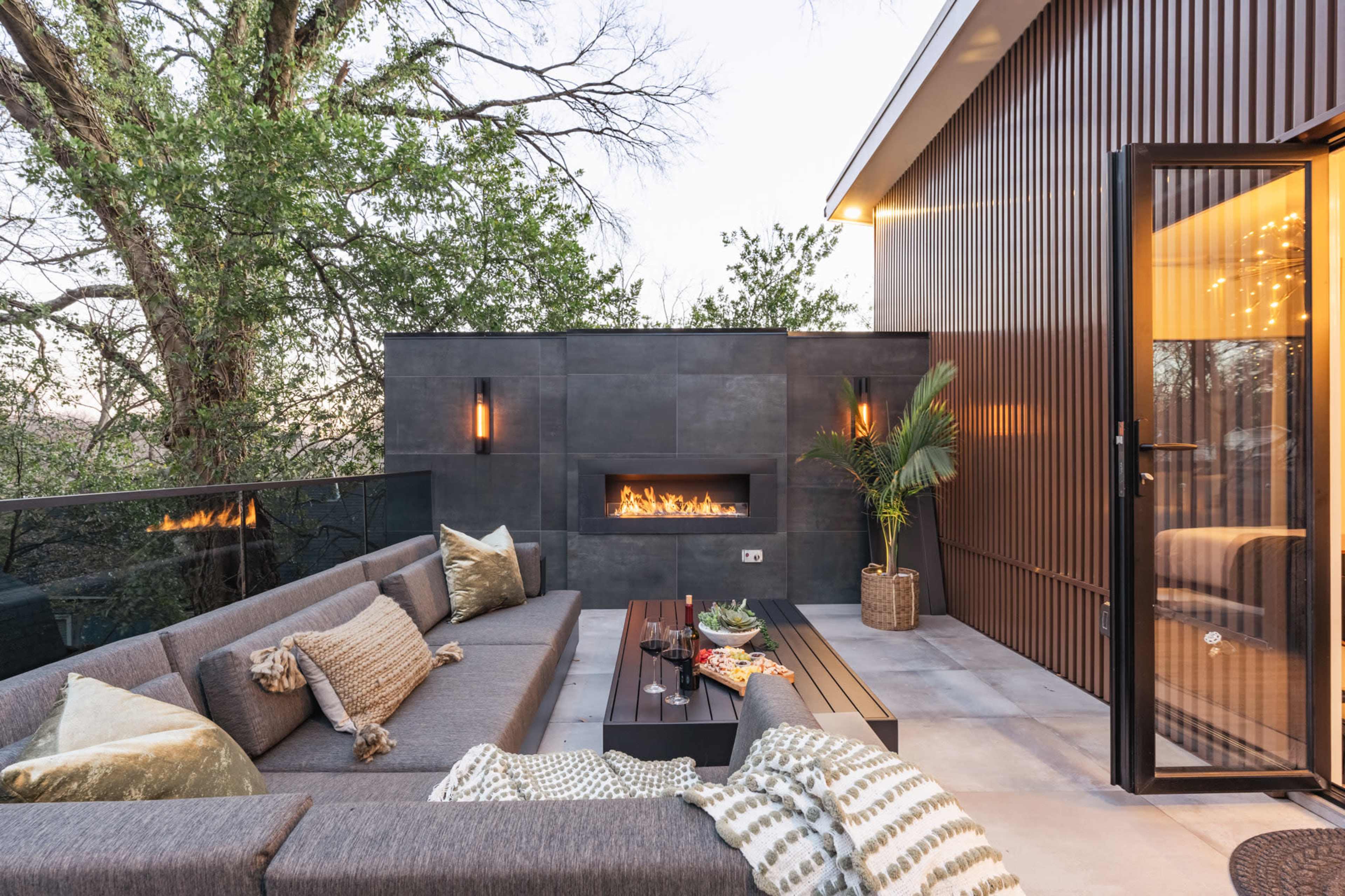 A modern outdoor patio features a sleek sectional sofa, a coffee table, and a built-in fireplace surrounded by greenery and a glass railing.