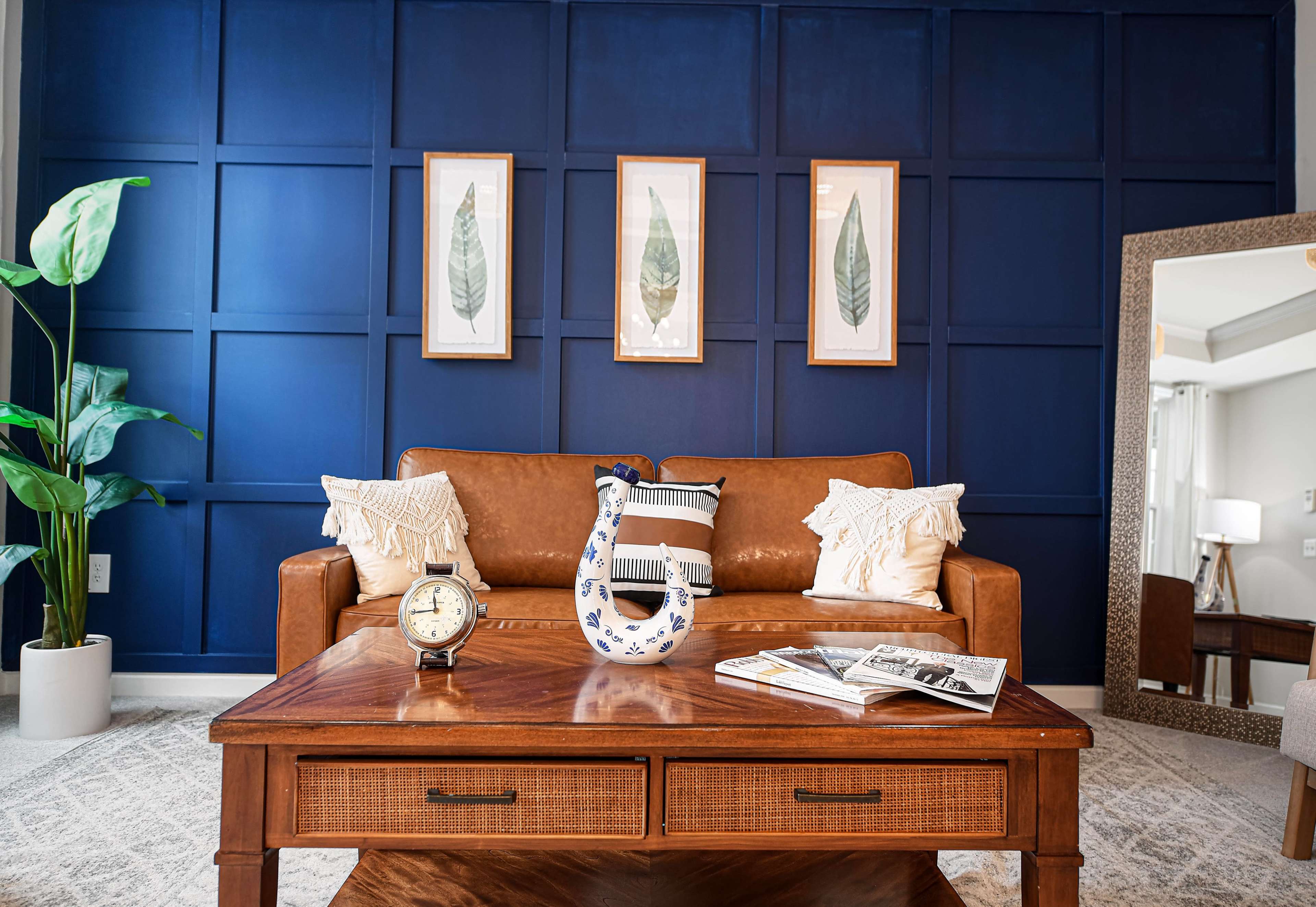 A living room with a brown leather sofa, a wooden coffee table, decorative vase, and framed botanical prints on a dark blue wall.