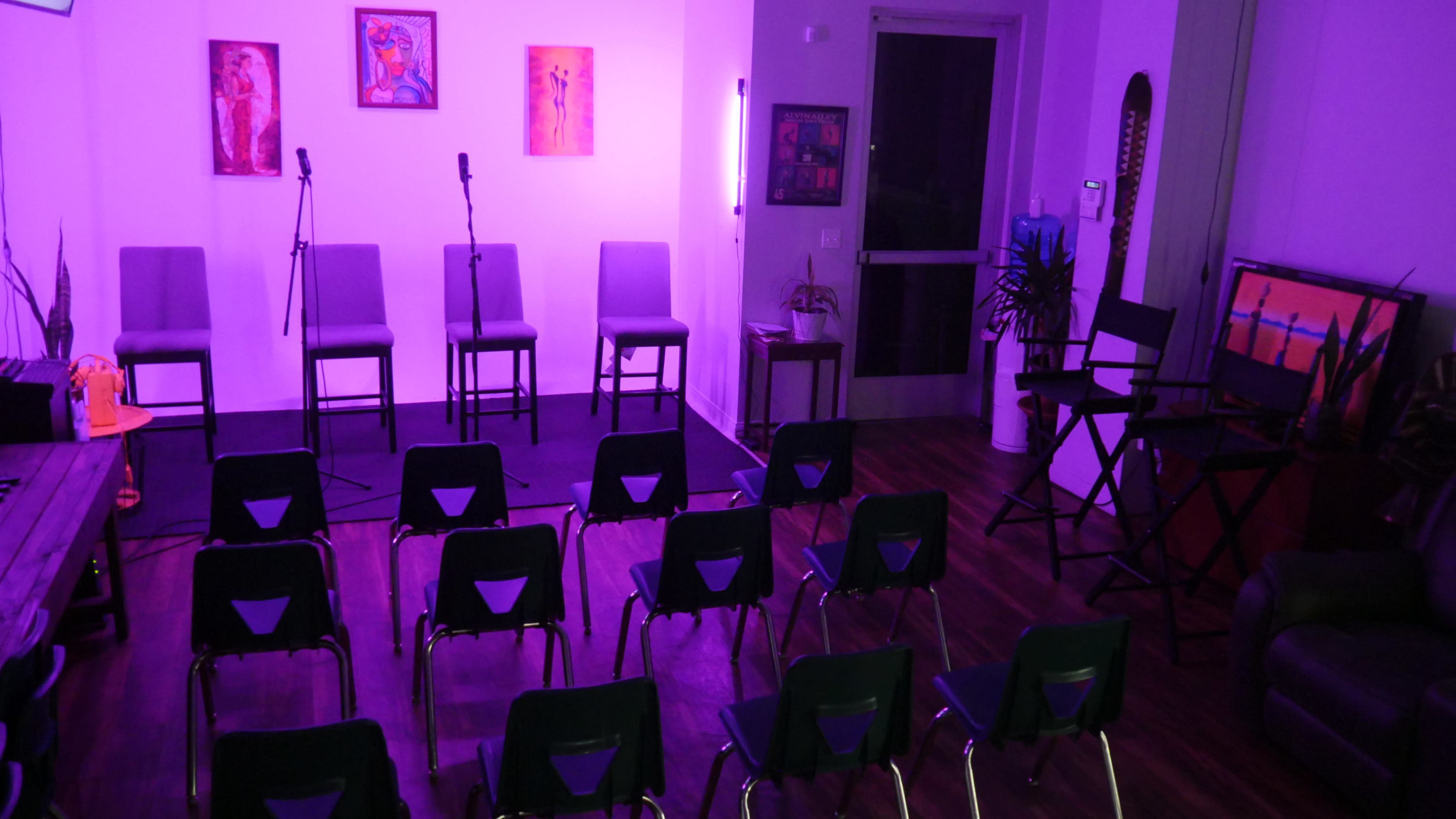 A small performance space with three empty chairs on a stage surrounded by rows of folding chairs, all illuminated by purple lighting.