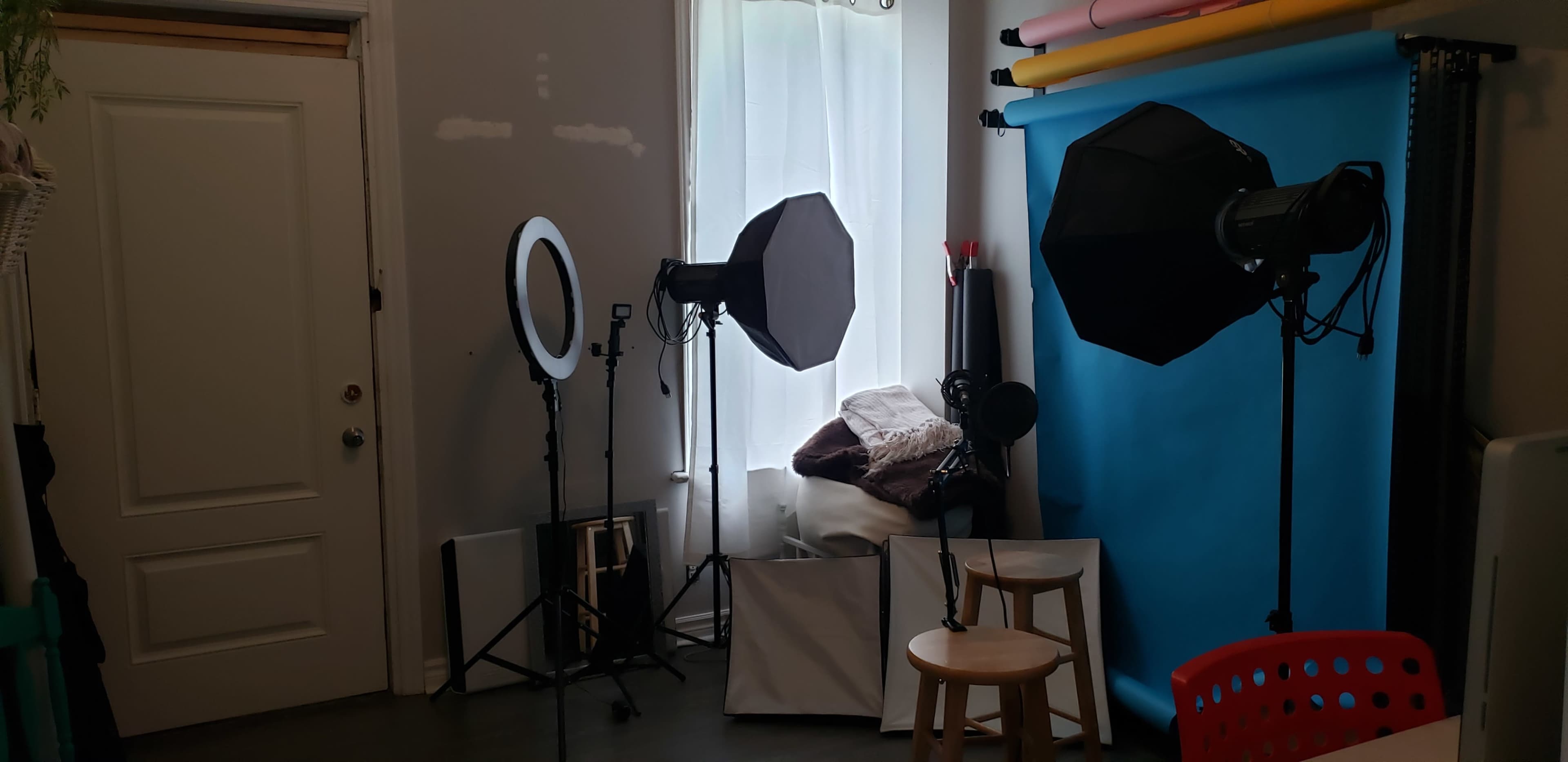 The image shows a photography setup in a room with lighting equipment, a backdrop, and a few chairs arranged near a window.