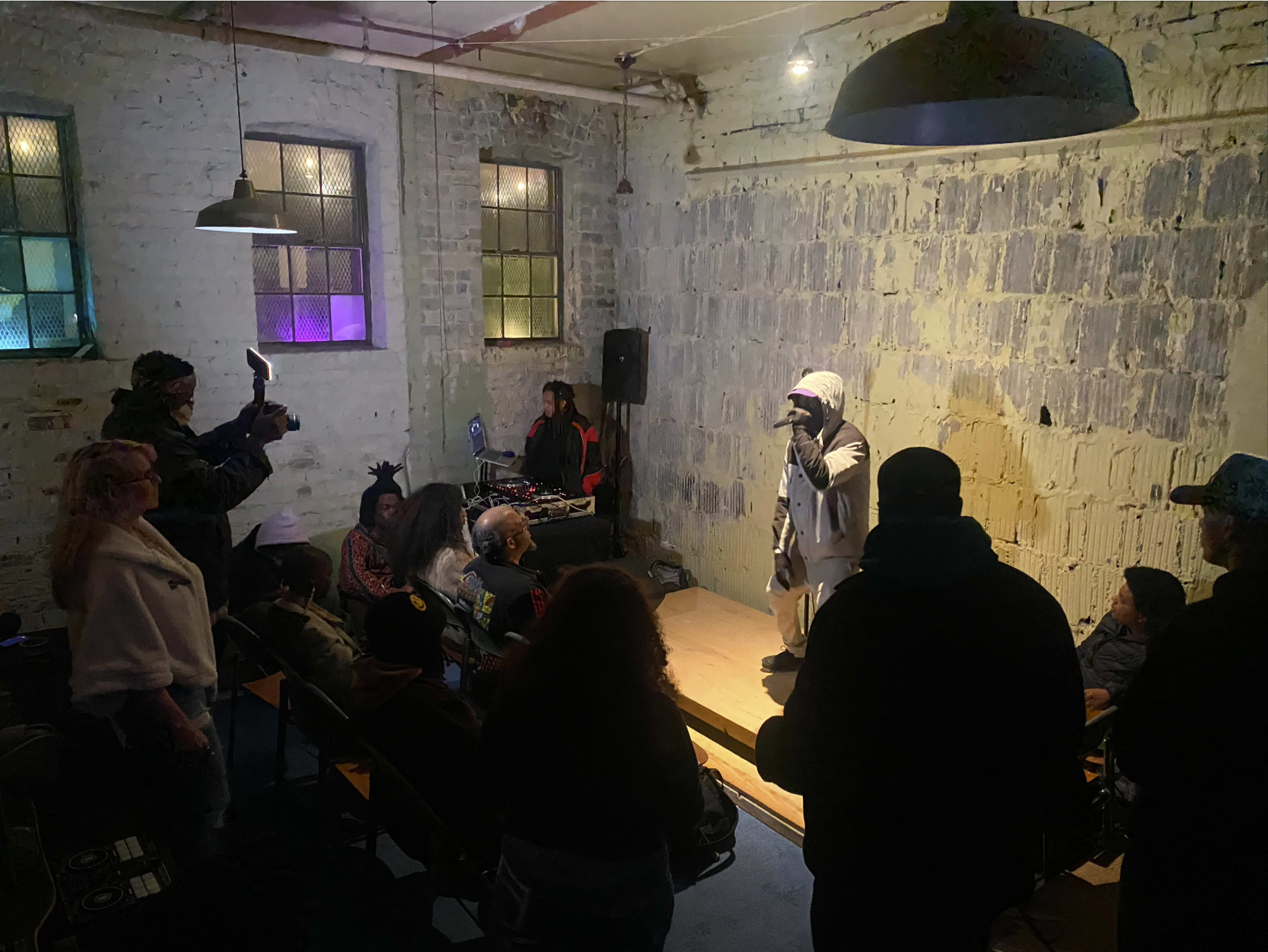 A performer is on stage in a small, dimly lit venue while an audience watches and a cameraman records the scene.
