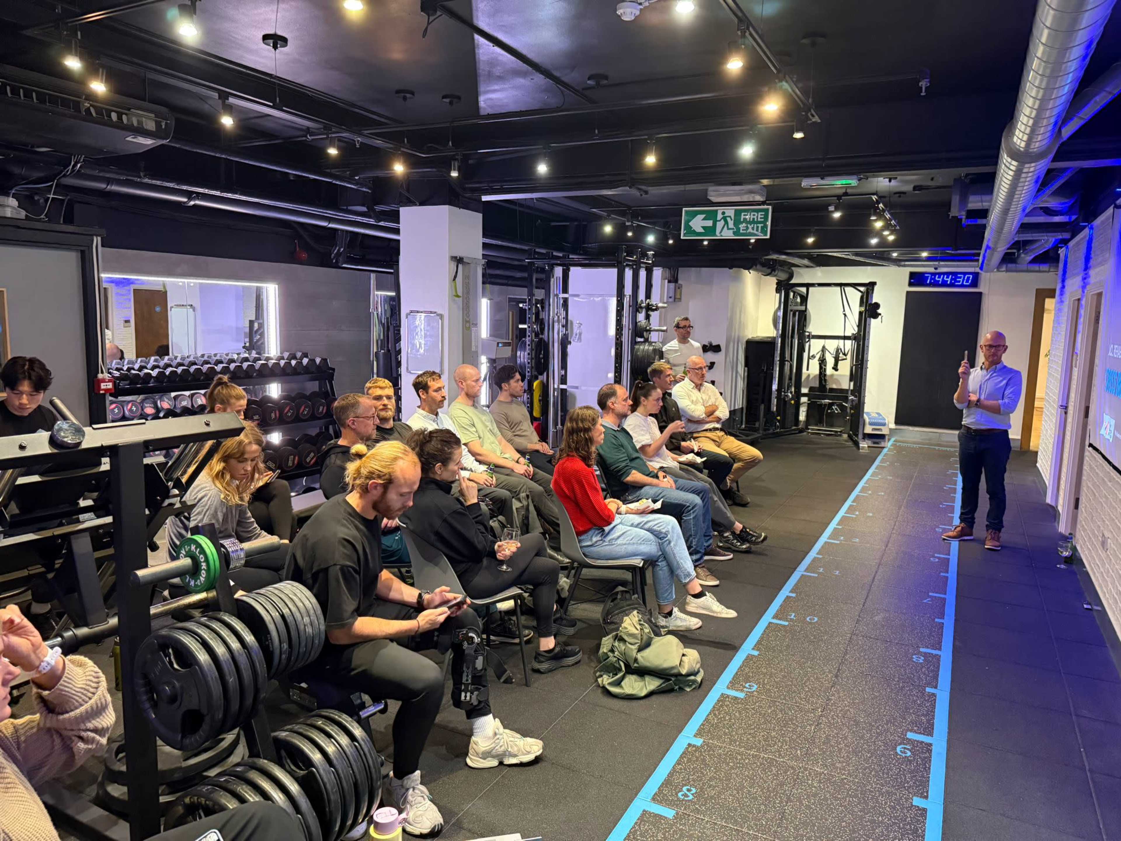 A group of people is seated in a gym with weightlifting equipment, while a presenter stands at the front, addressing the audience.