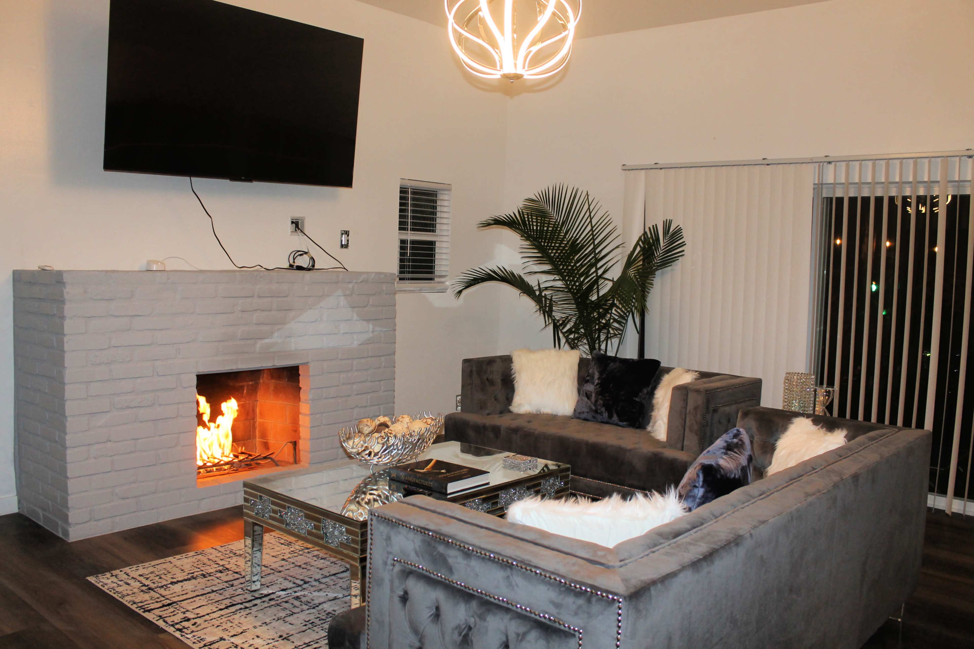 A modern living room features a gray sectional sofa, a glass coffee table, a brick fireplace with a flame, and a wall-mounted television, complemented by a tall indoor plant and vertical blinds.