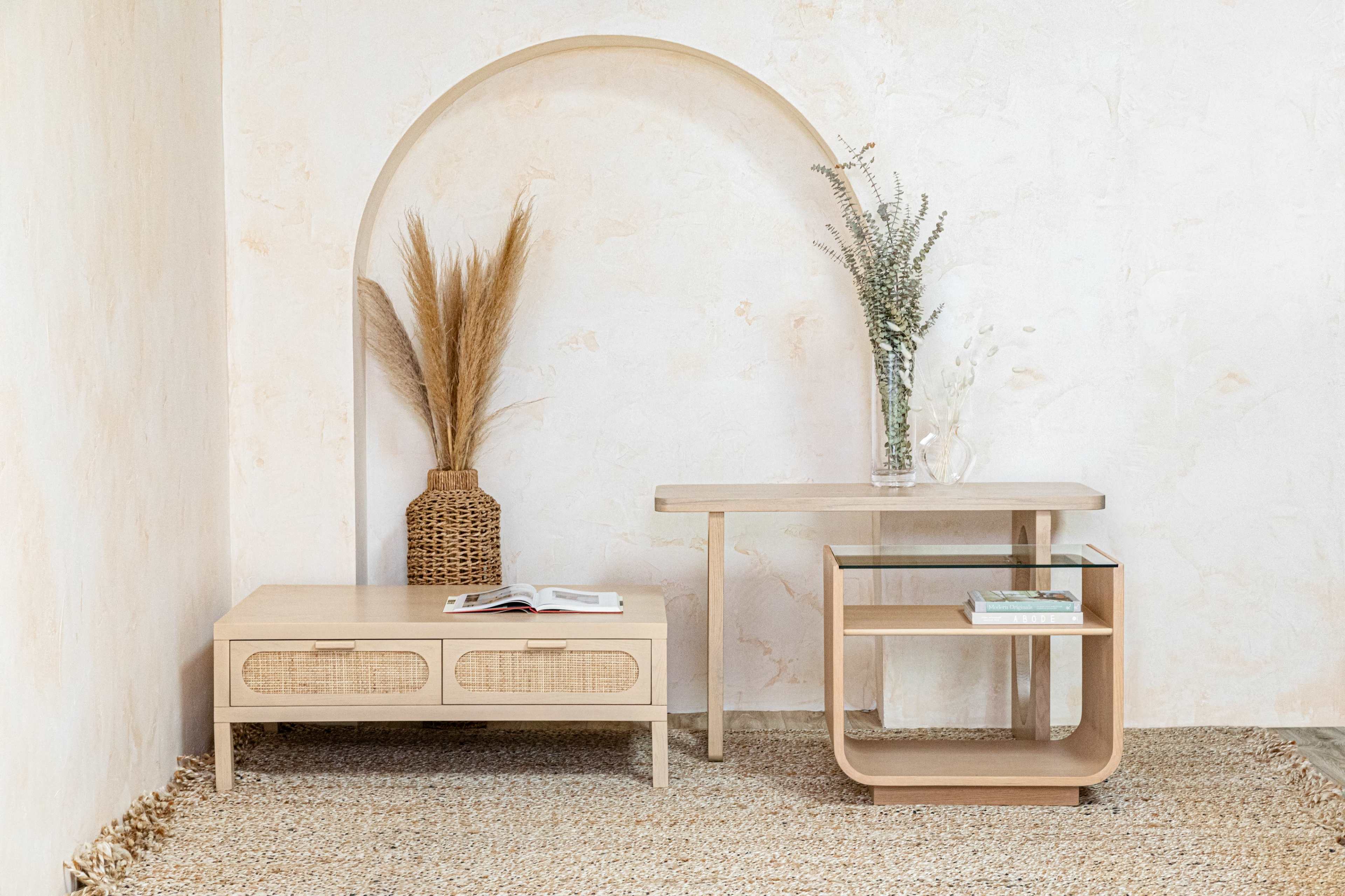 A light-colored room features a minimalist setup with a wooden table, a woven basket holding pampas grass, and a shelf, all positioned on a textured rug.