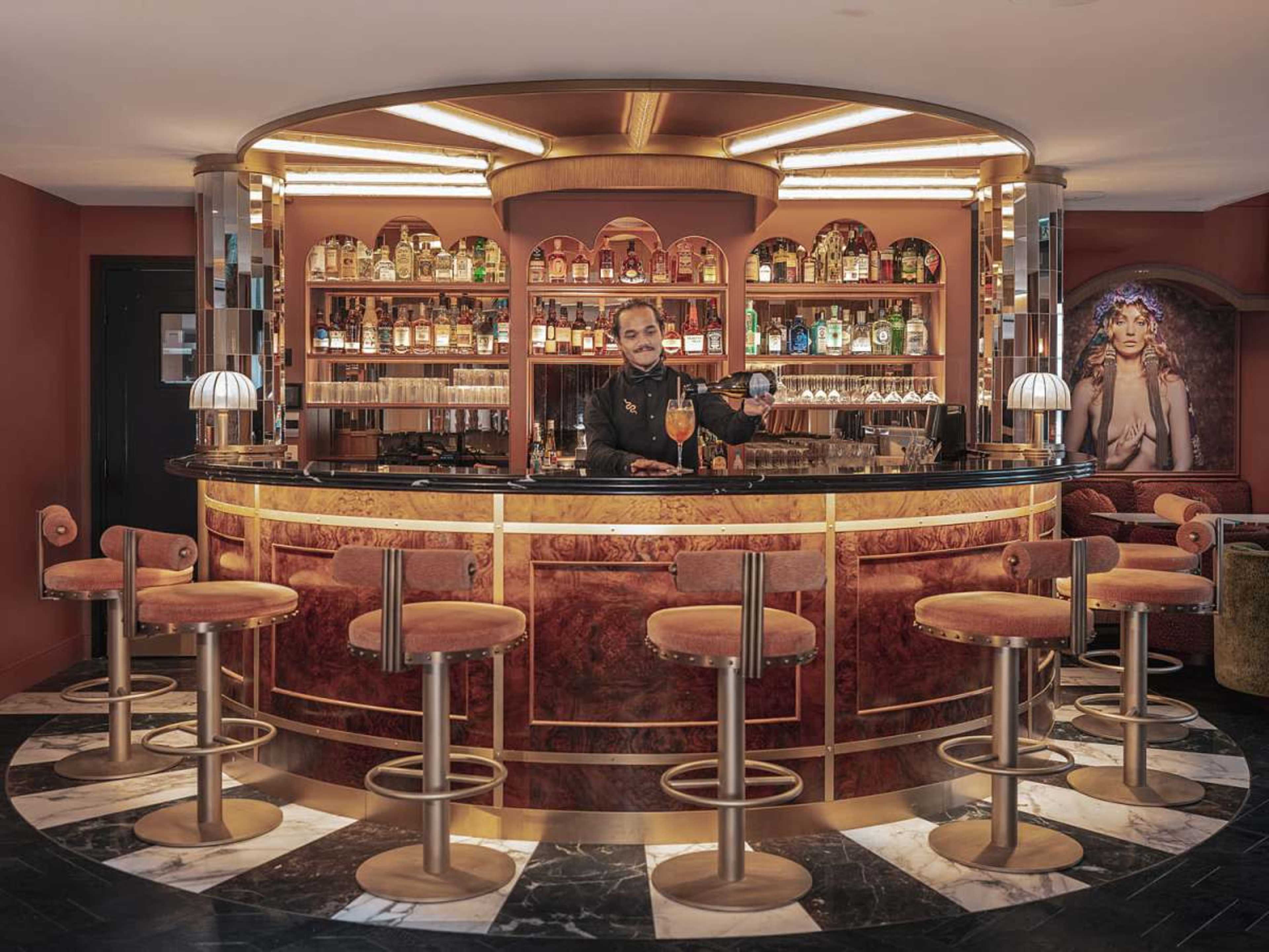 A bartender serves a colorful drink at an elegantly designed bar featuring illuminated shelves stocked with various bottles.