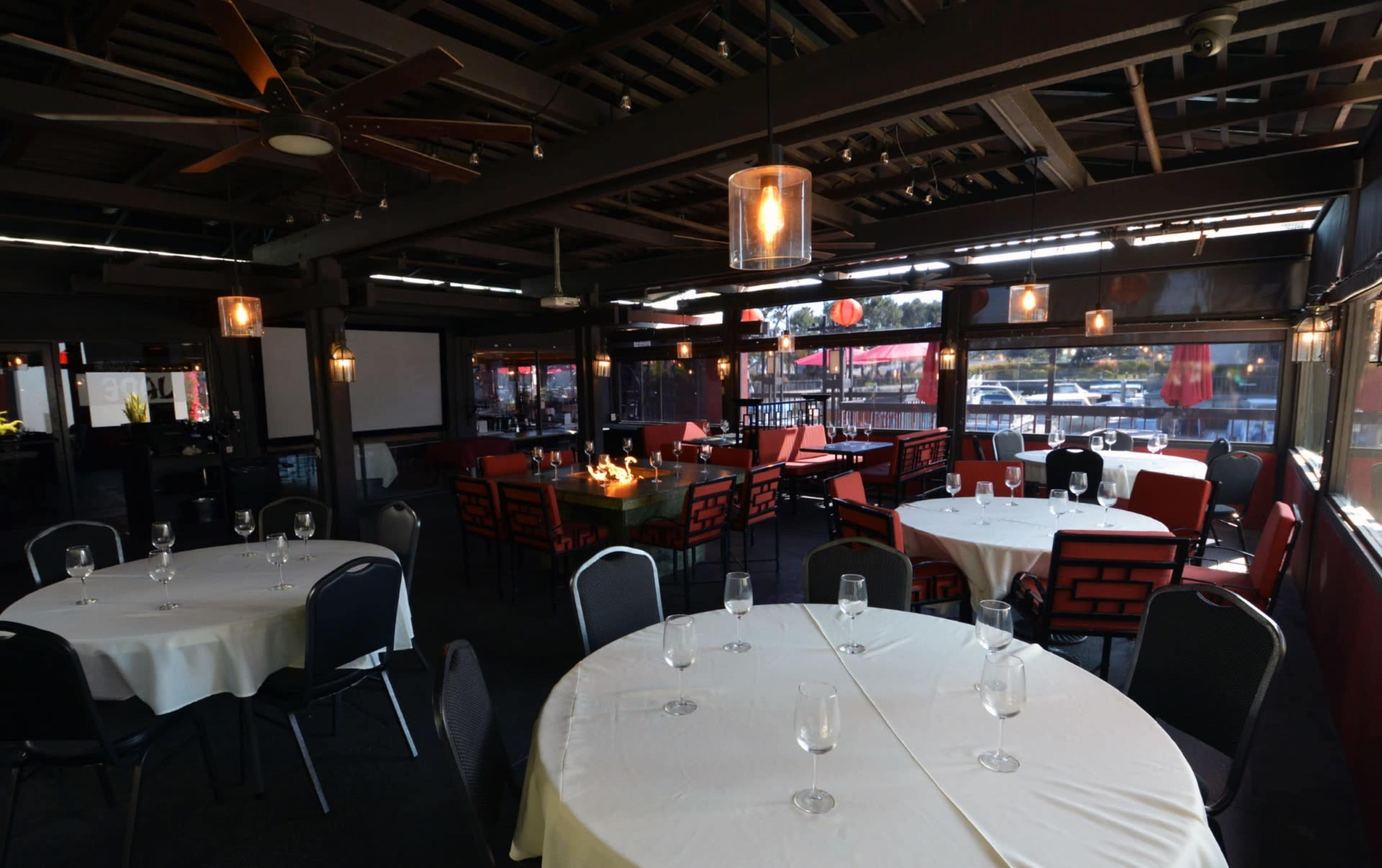 An indoor dining area with tables set for meals, featuring round white tablecloths and black chairs, surrounded by large windows.
