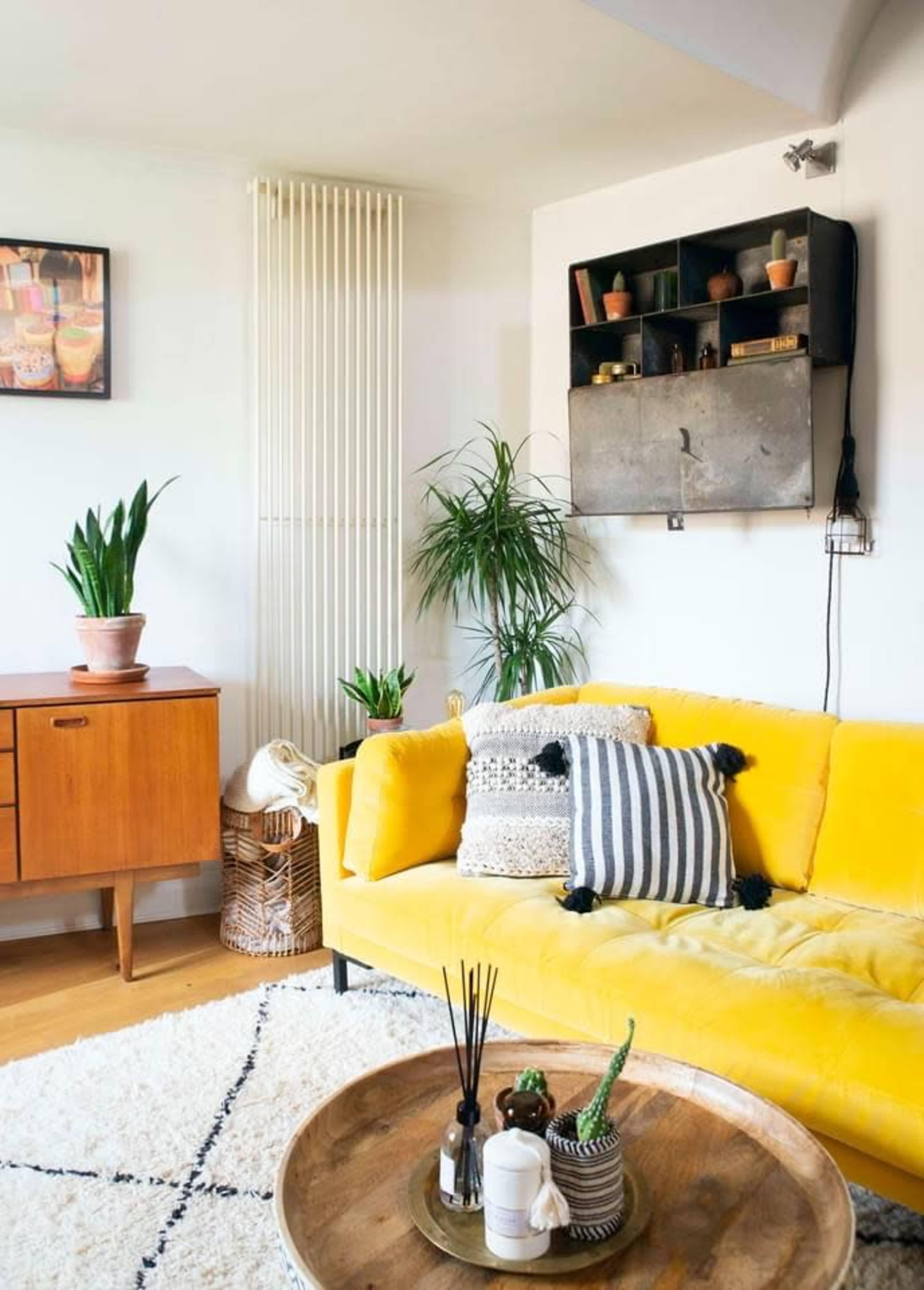The living room features a yellow couch, a wooden cabinet, and plants, complemented by a round wooden coffee table on a textured rug.