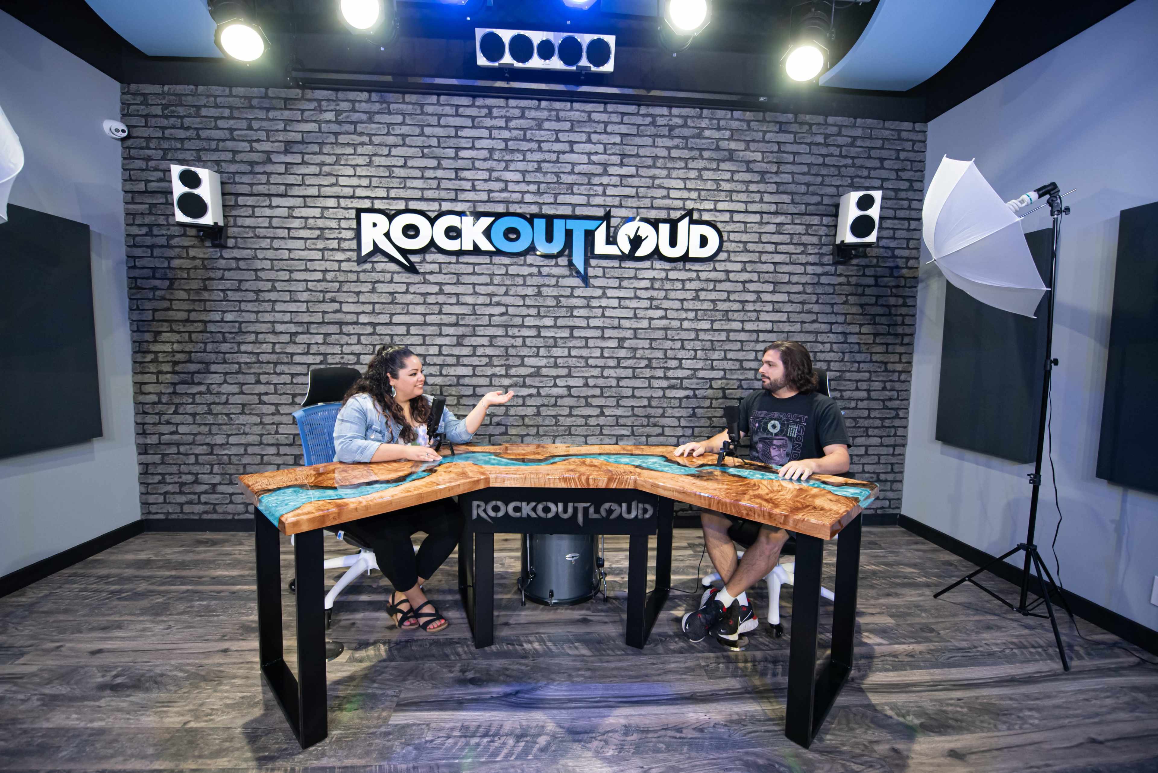 Two people are seated at a uniquely designed wooden table in a studio with a brick wall backdrop, discussing in front of camera equipment and lighting.