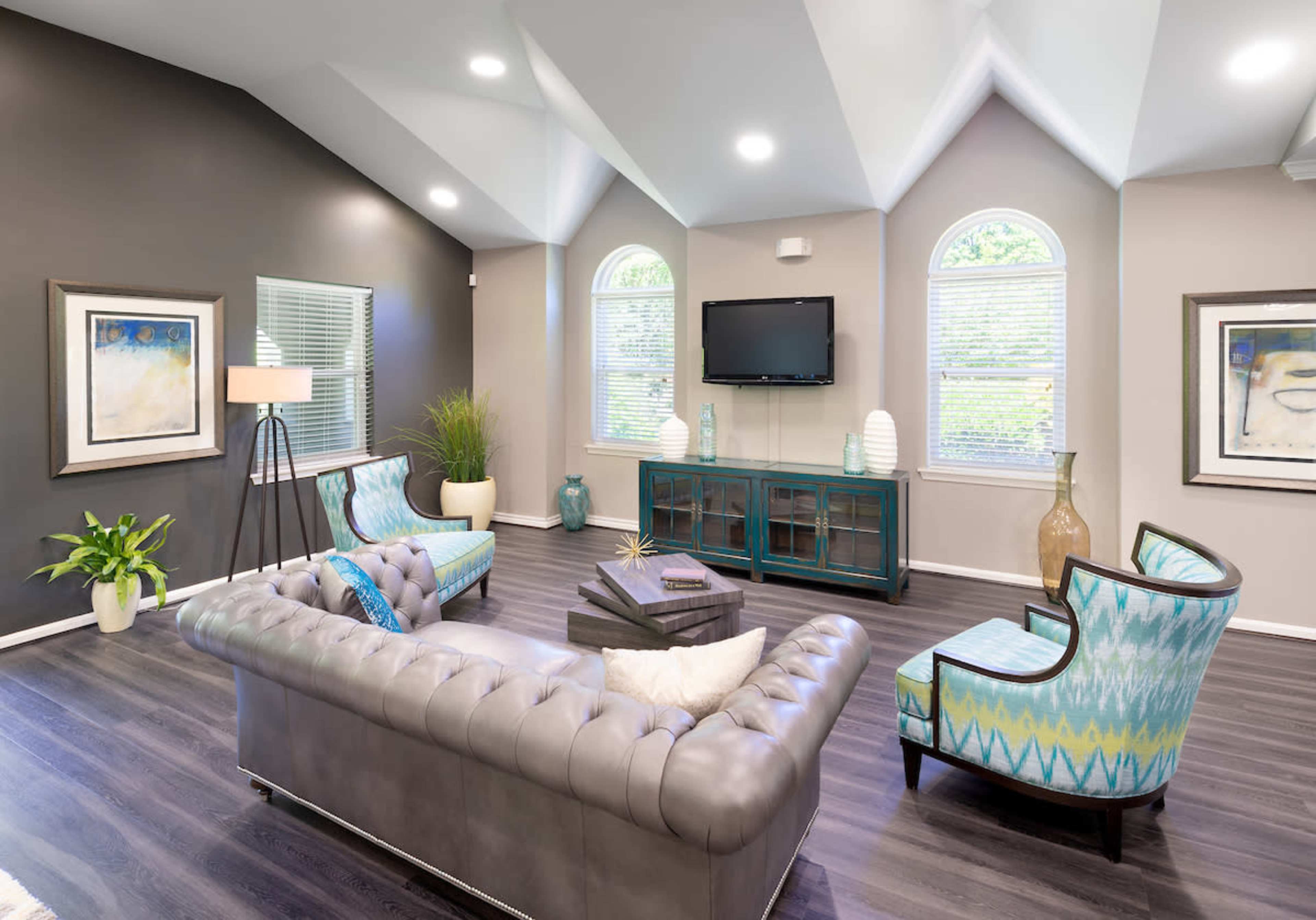 The image shows a modern living room with a gray leather couch, two patterned chairs, a television mounted on the wall, and large windows allowing natural light to enter.