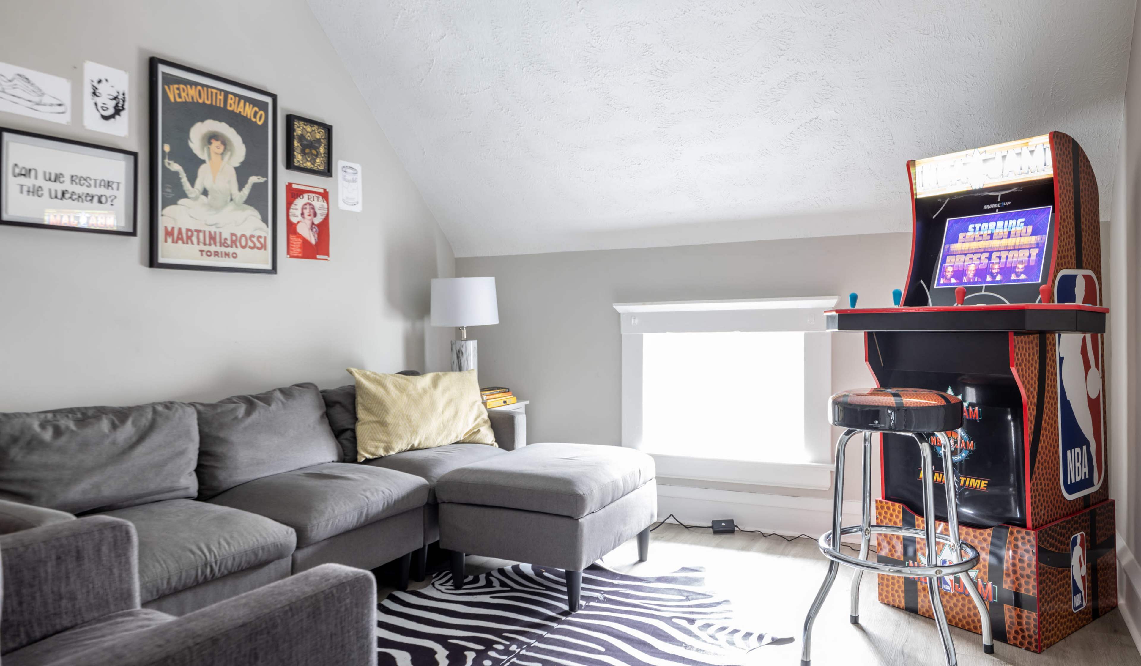 A cozy living room with a gray sectional sofa, a small barstool next to an arcade game, and several framed posters on the wall.