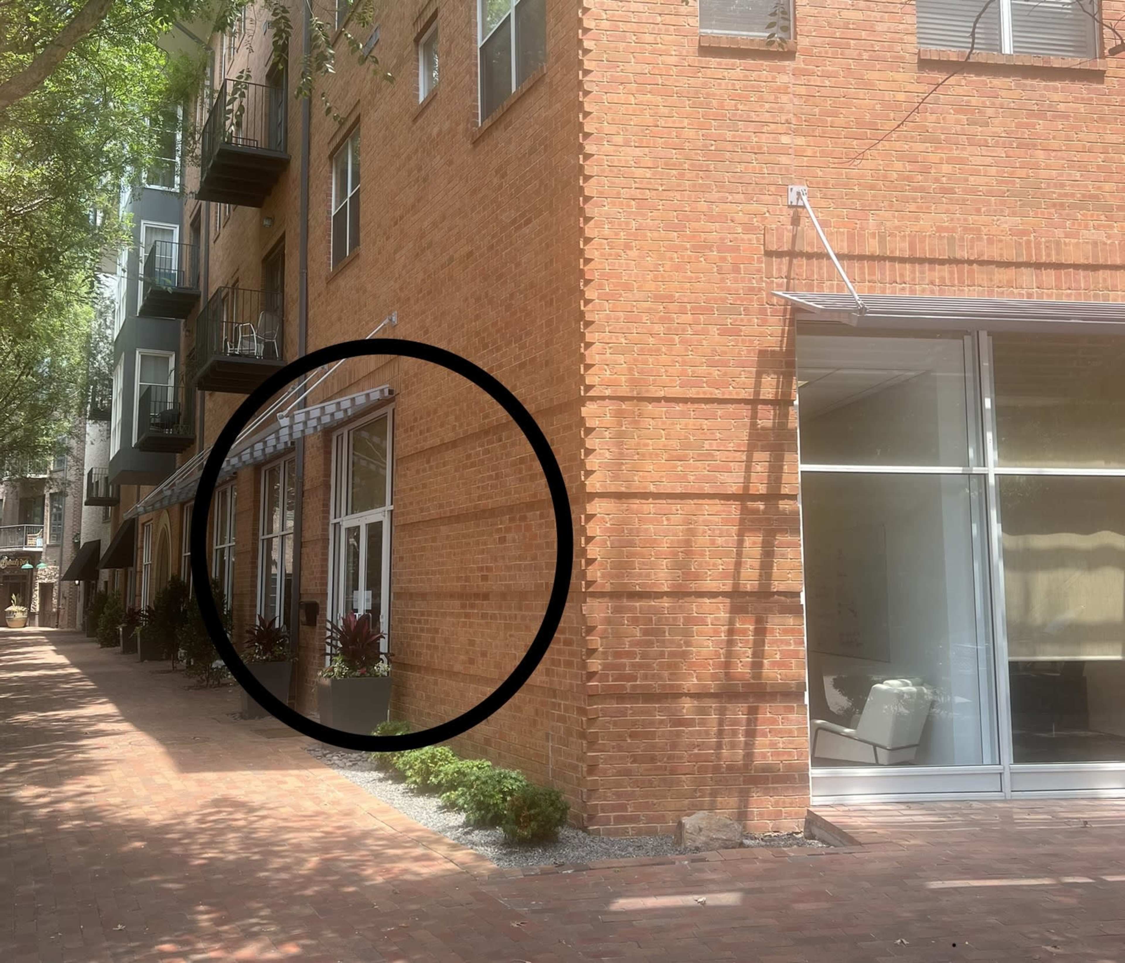 The image shows a brick building with large windows and a pathway beside it, flanked by greenery and additional structures.
